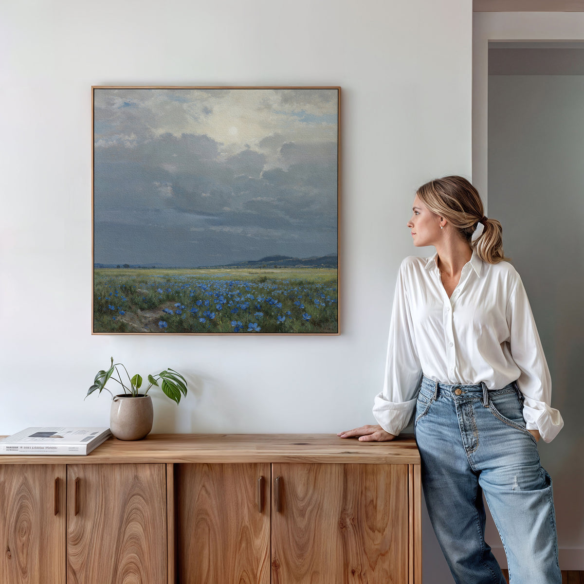 A woman in a white blouse and blue jeans leans against a wooden sideboard, admiring the Cornflower Fields Canvas Art - Square, featuring a countryside landscape with cornflower fields and a cloudy sky. A plant and books sit on the sideboard.