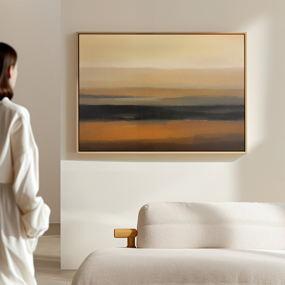 A woman in a white blouse stands in a bright, minimal room, admiring the Desert Mirage Canvas Art - Horizontal with warm earth tones and dark stripes, displayed above a cream-colored sofa.