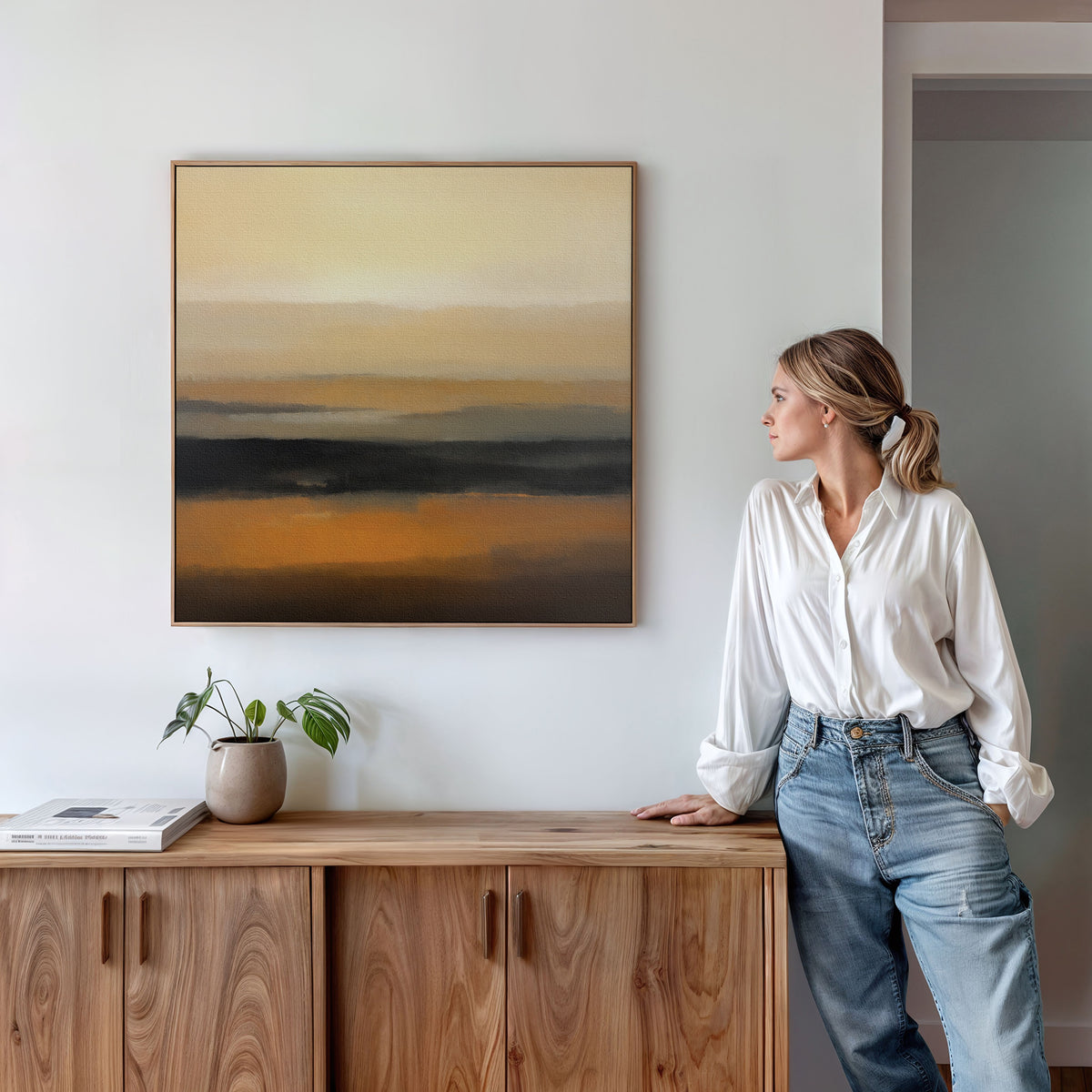 A woman in a white blouse and jeans stands by a wooden cabinet, admiring the Desert Mirage Canvas Art - Square. A potted plant and a book sit on the cabinet beneath the warm, earth-toned Nordic Park-inspired artwork.