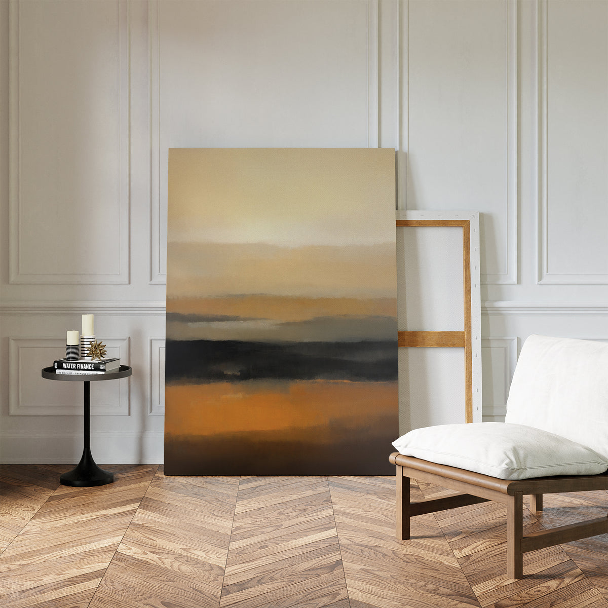 The Desert Mirage Canvas Art - Vertical, featuring abstract earth tones, leans against a white paneled wall in a modern room with an empty frame, a white cushioned chair, and a small black table holding candles and books on hardwood floors.