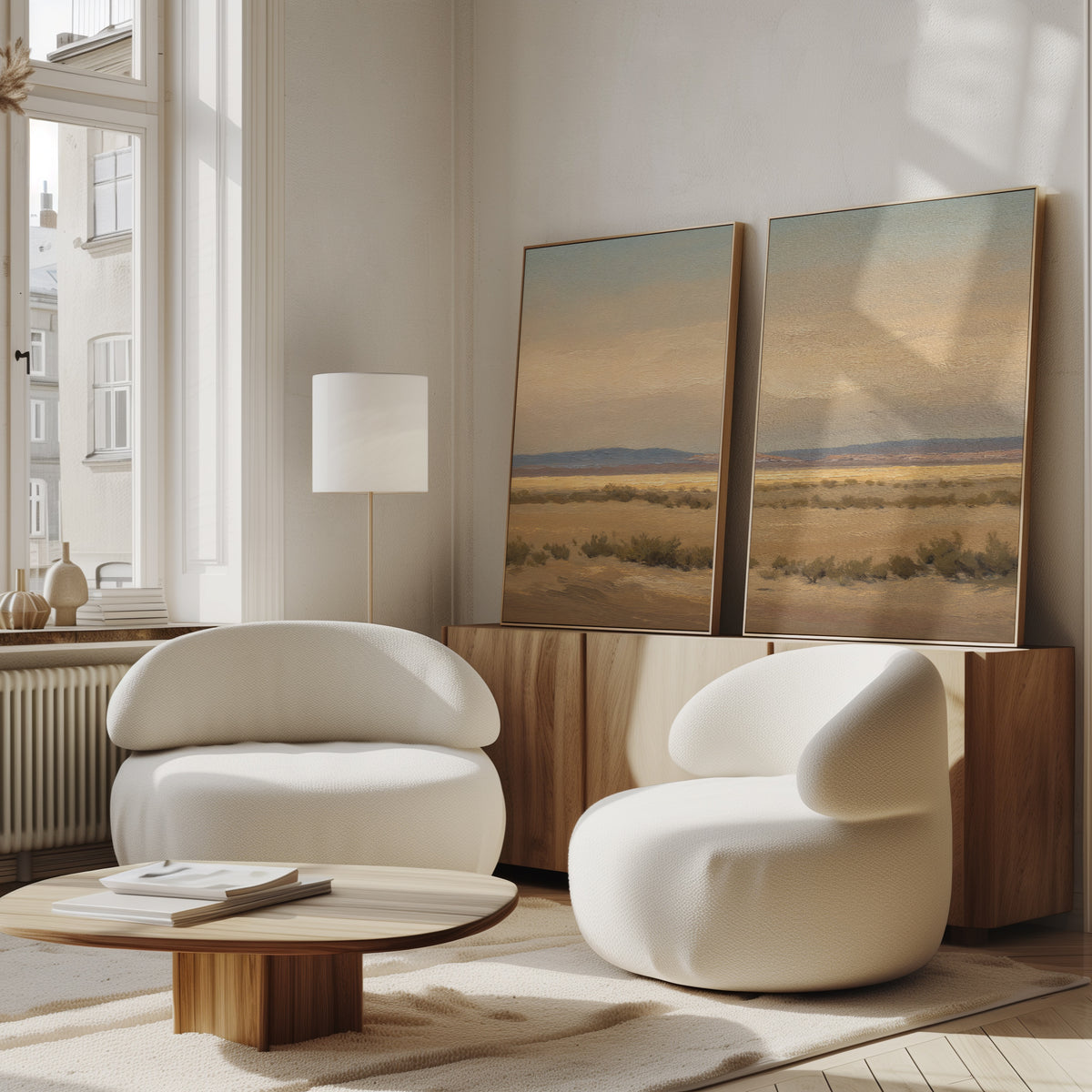 A modern living room features two white rounded armchairs, a low wooden coffee table, and a sideboard topped with the "Desert Solitude - Set of 2" large landscape canvas prints. Sunlight through tall windows creates a warm, inviting vibe.