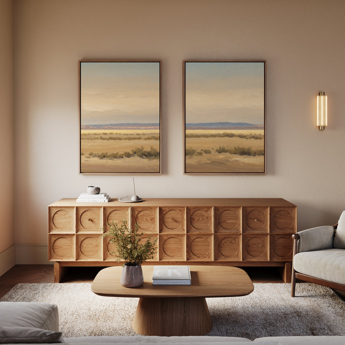 A cozy living room featuring the Desert Solitude - Set of 2 southwestern landscape wall art, a wooden sideboard, a round coffee table with a vase and book, a light gray armchair, and warm neutral decor.