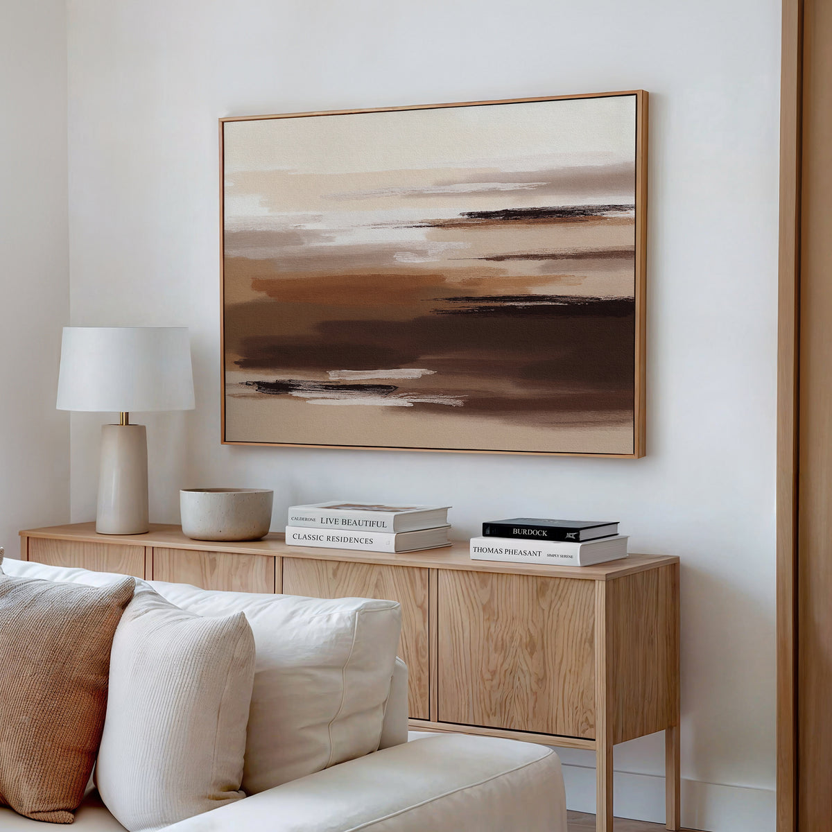 Minimalist living room corner featuring a wooden sideboard, books, white table lamp, ceramic bowl, light sofa, and the Desert Winds Canvas Art - Horizontal in warm brown, beige, and cream tones displayed on the wall.