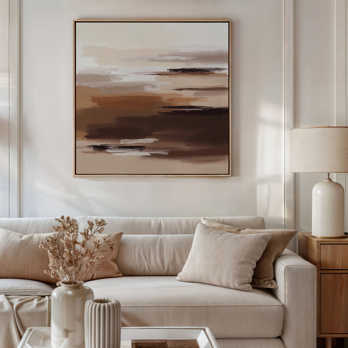 A modern living room with a beige sofa, neutral cushions, and a wooden side table with a cream lamp. Above, the Desert Winds Canvas Art - Square hangs as earth tone wall art. Dried flowers in a vase sit on the coffee table.