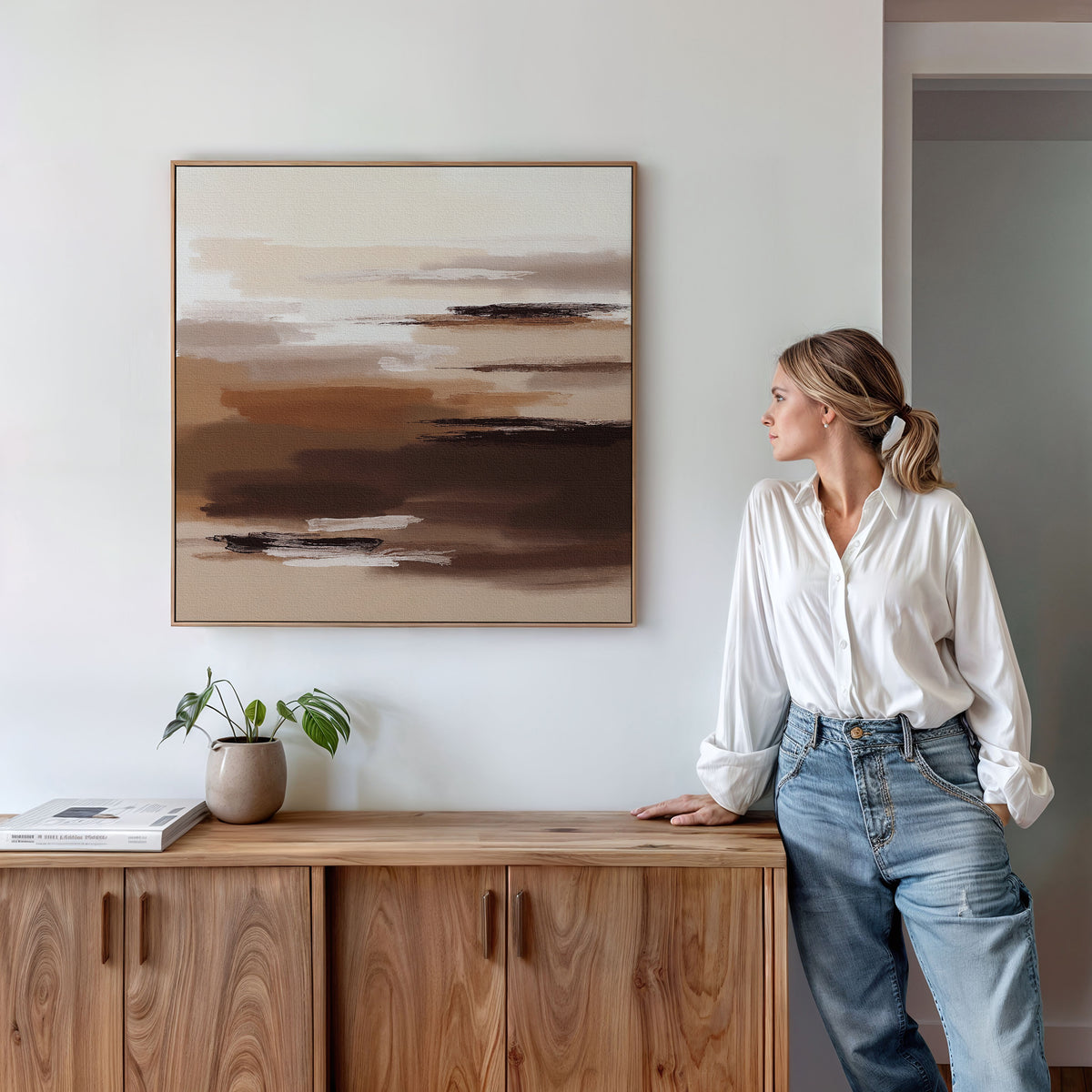 A woman in a white blouse and blue jeans leans against a wooden cabinet, admiring the Desert Winds Canvas Art - Square on the wall. A small potted plant and a book are placed on the cabinet.
