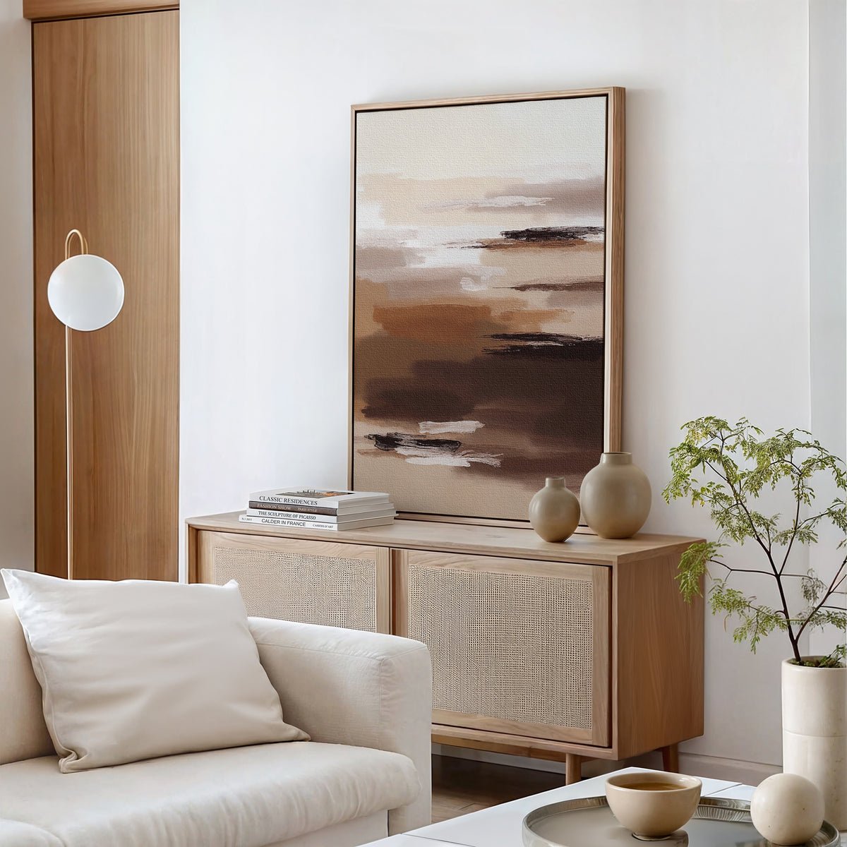 A minimalist living room with a light wooden sideboard, Desert Winds Canvas Art - Vertical, neutral vases, stacked books, a white sofa with a cushion, a small potted plant, and a round hanging light—perfect for Nordic Park style.