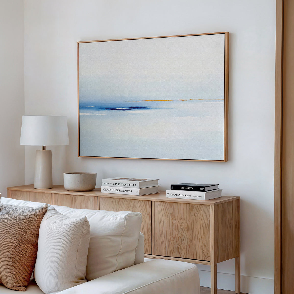 A light, modern living room featuring a wooden sideboard, white lamp, stacked books, a bowl, and the Distant Archipelago Canvas Art - Horizontal in soft blue and beige tones hanging on the white wall above.