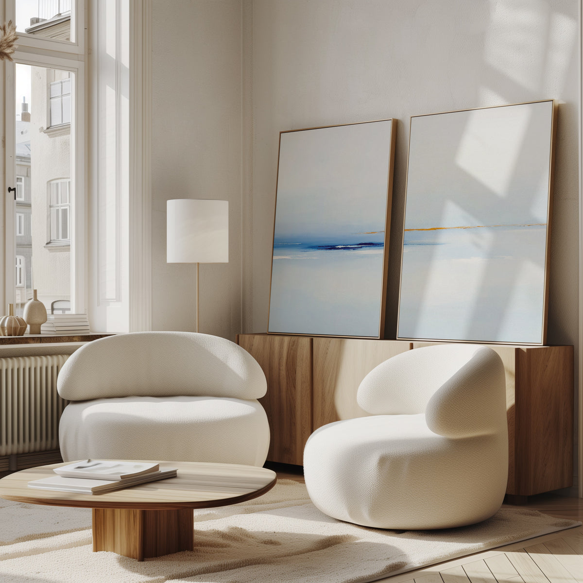 A modern living room with two rounded white armchairs, a low wooden coffee table, and the Distant Archipelago - Set Of 2 wall art displayed on a wooden sideboard near tall windows that fill the space with natural light.