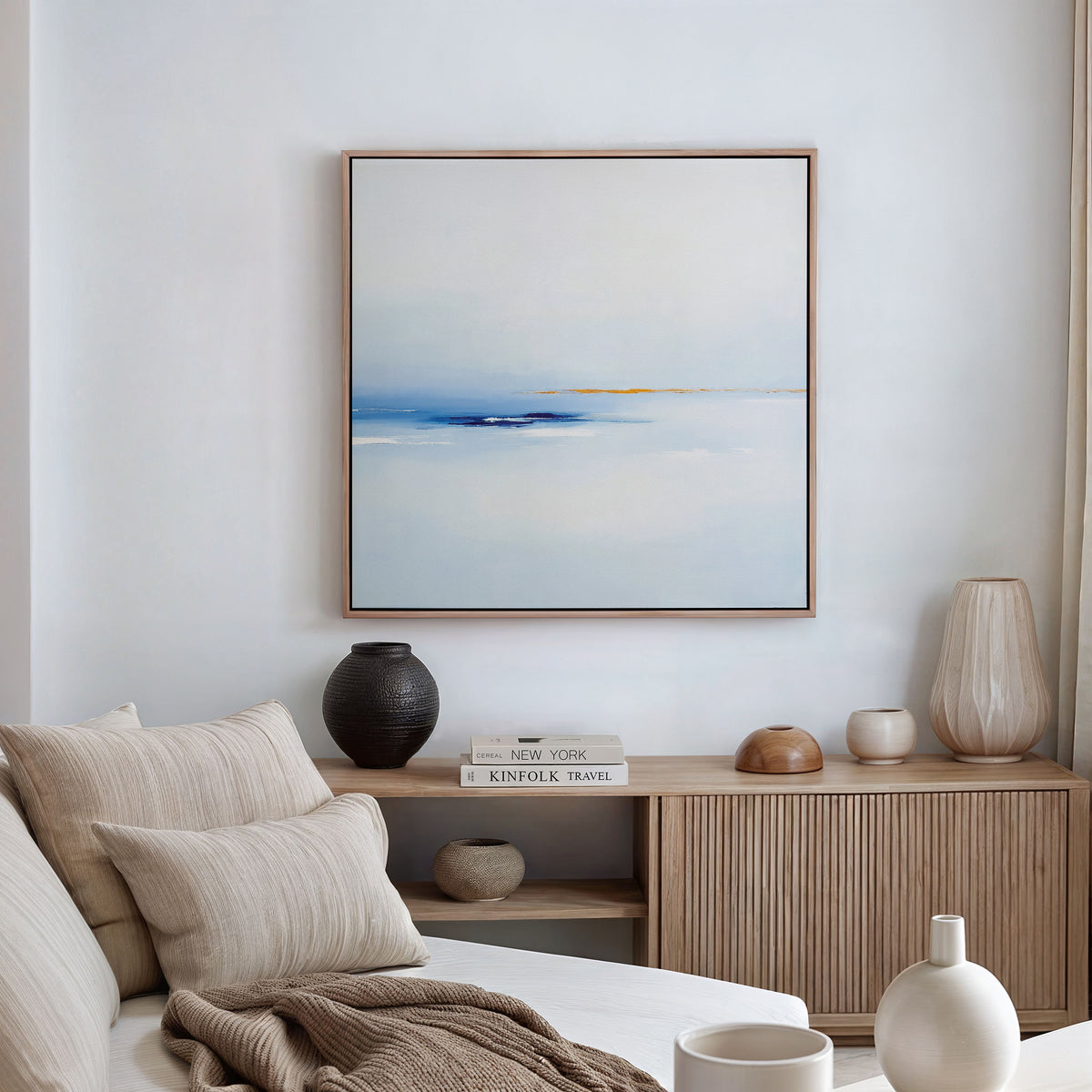 A modern, minimalist living room features a beige sofa, neutral pillows, wooden sideboard, decorative vases, stacked books, and the Distant Archipelago Canvas Art - Square in striking blue and white tones displayed on the wall.