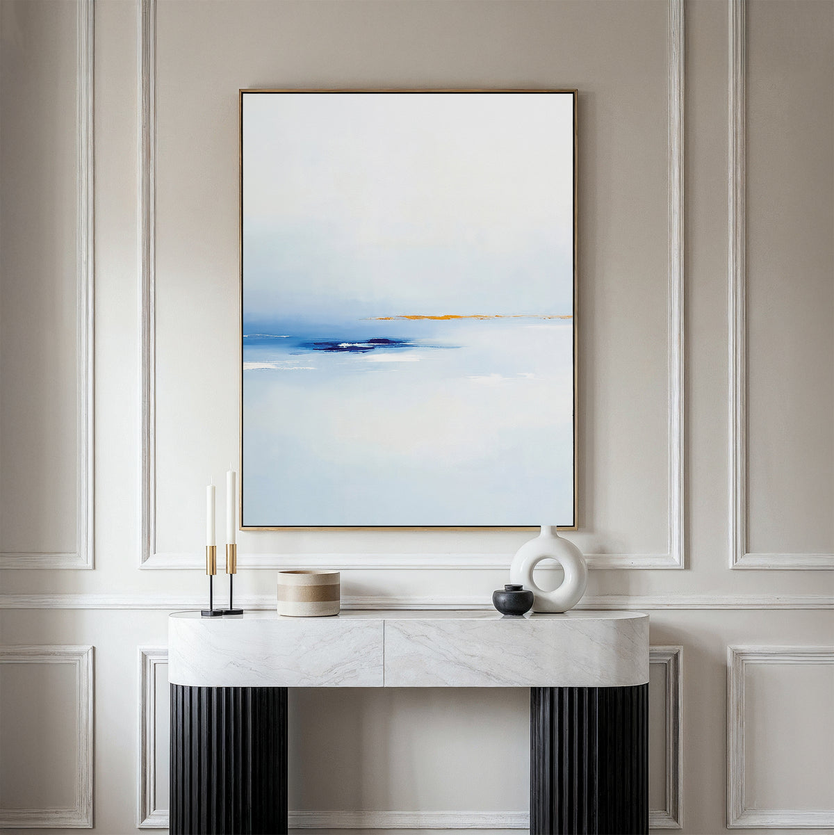 A minimalist living space with a white paneled wall displays the Distant Archipelago Canvas Art - Vertical in soft blue and white above a white marble console table adorned with modern vases and candles.