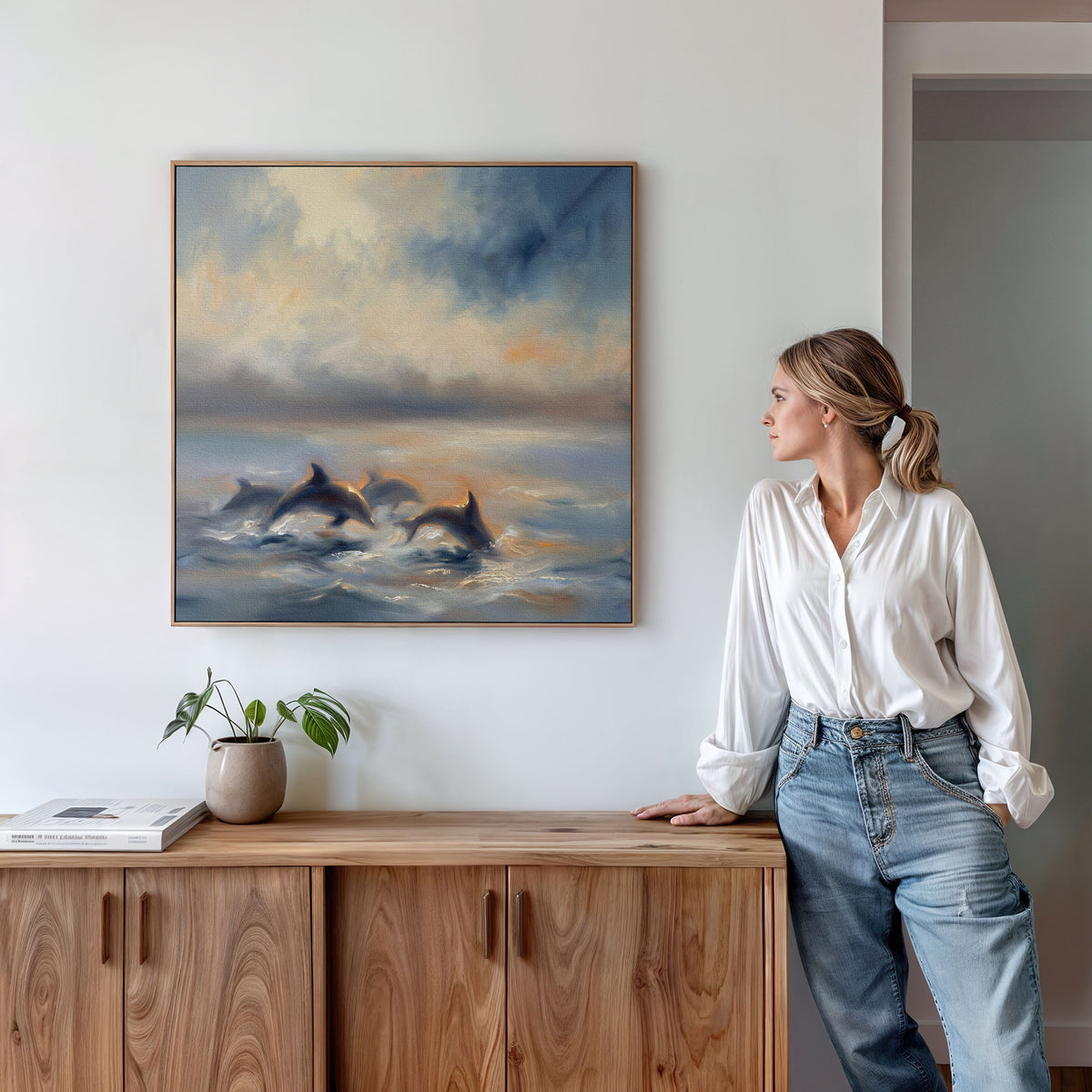 A woman in a white blouse and jeans stands by a wooden cabinet, admiring the "Dolphin Pod at Sunset Canvas Art - Square" that hangs on the white wall above, showcasing dolphins leaping in an ocean sunset.