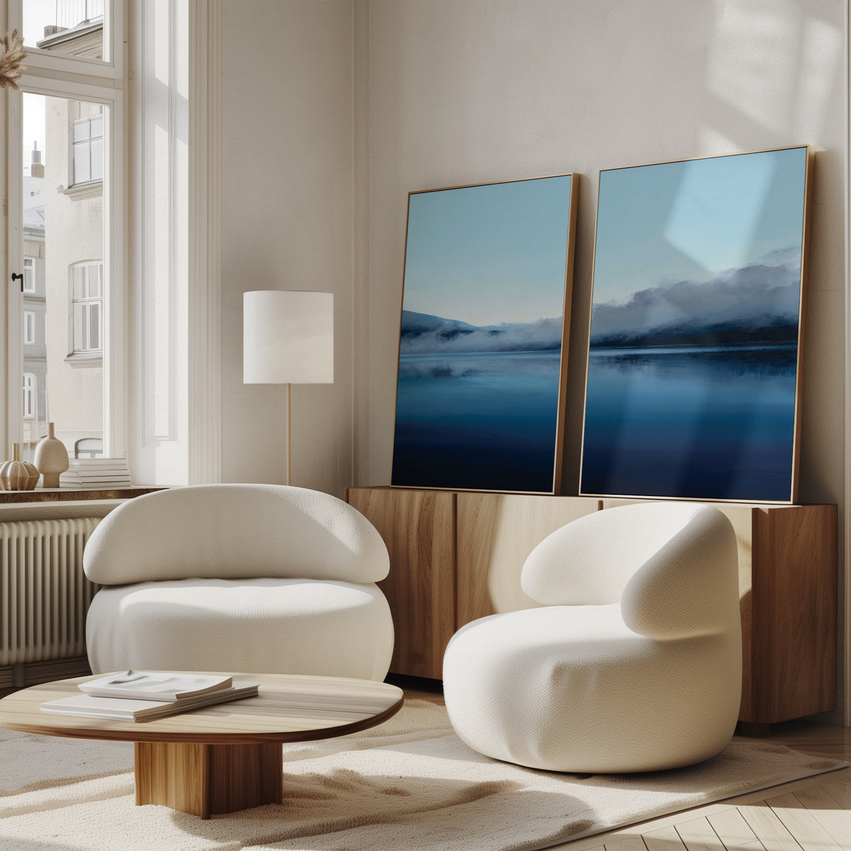 Two modern white armchairs and a low wooden table sit in a bright room with large windows. On the sideboard, two framed "Fjord Mist - Set Of 2" prints—one featuring blue waters—capture the essence of Nordic Park.