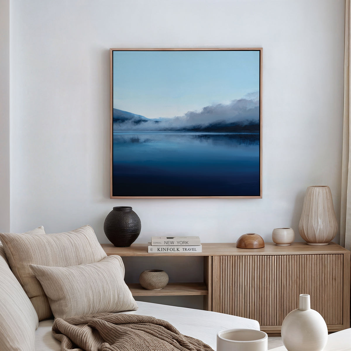 A modern, minimalist living room features a beige sofa, striped pillows, a light wood sideboard, and the Fjord Mist Canvas Art - Square—a serene Nordic landscape print of a calm blue lake with mountains—hanging on the white wall.