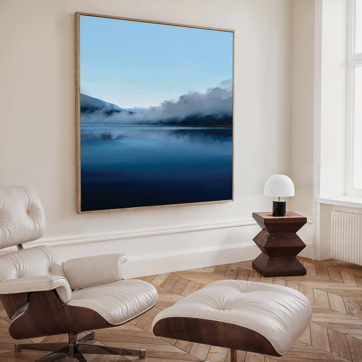 A modern living room with a white leather lounge chair, matching ottoman, geometric wooden side table with a round lamp, and the Fjord Mist Canvas Art - Square featuring misty mountains over a calm blue lake on the wall.