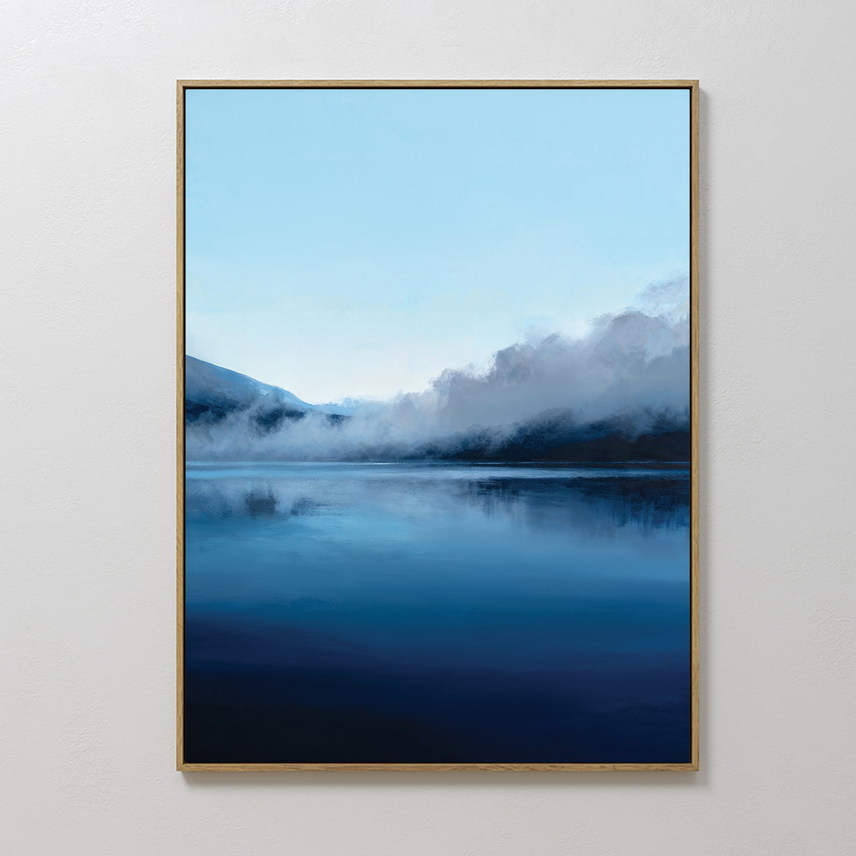 The Fjord Mist Canvas Art - Vertical features a serene lake with misty mountains reflected on calm blue water under a clear sky. A simple gold frame and white wall make this tranquil piece perfect for Nordic-inspired decor.