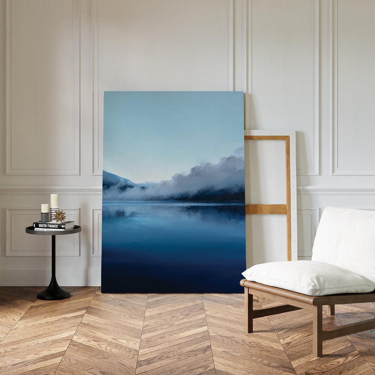 A minimalist room with herringbone floors, a white chair, and Nordic decor features a round black side table with candles and the Fjord Mist Canvas Art - Vertical propped against a white paneled wall.