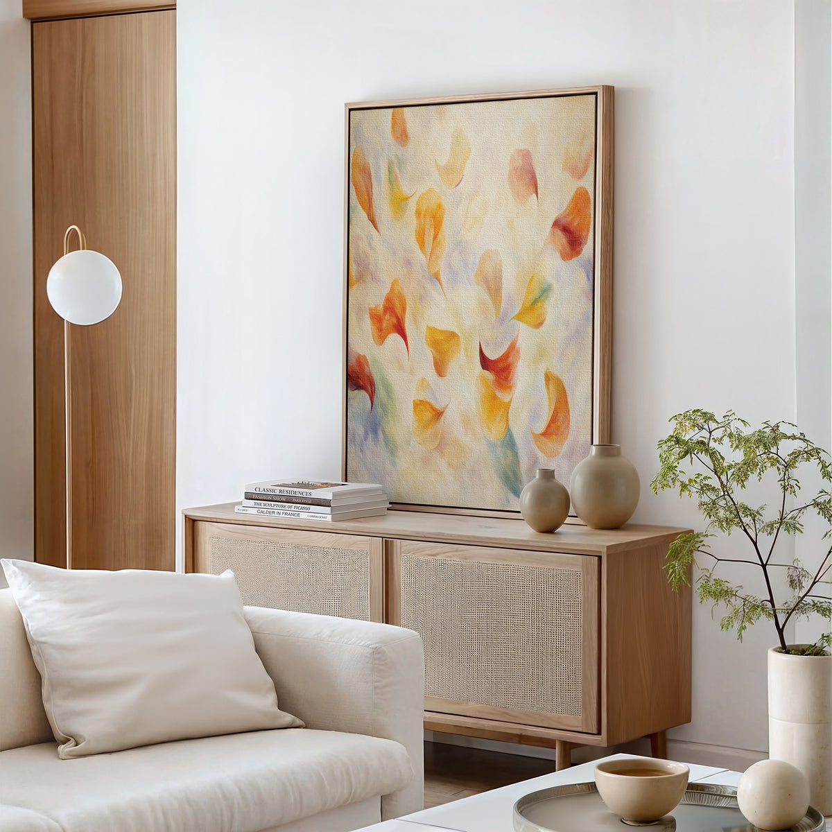 A modern living room with a neutral sofa, wooden sideboard, ceramics, and a potted plant features “Floating Petals Canvas Art - Vertical,” adding organic elegance in soft orange, yellow, and red tones to the wall.