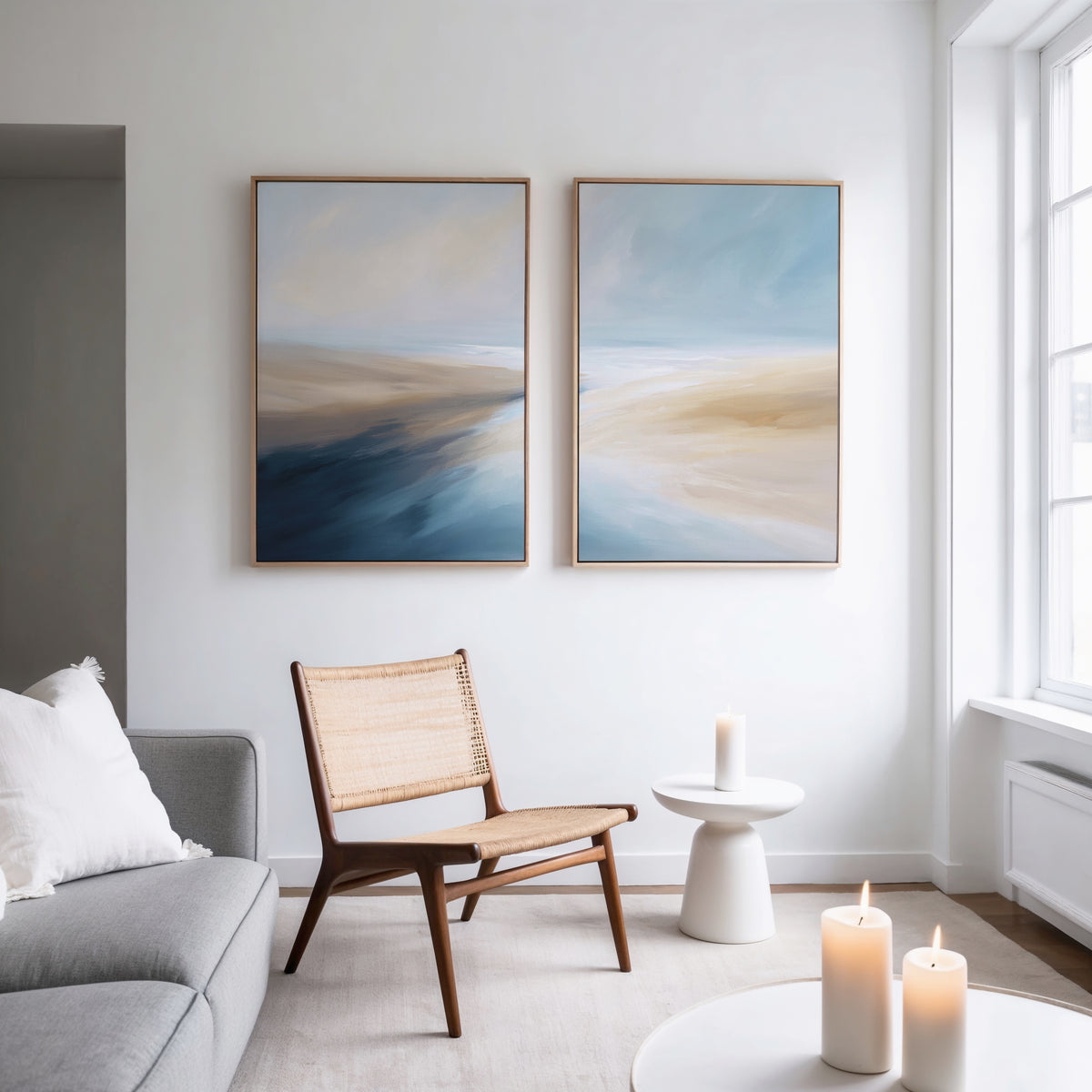 A modern living room with a light gray sofa, woven wooden chair, candles on sleek tables, and Fluid Motion - Set Of 2 wall art pieces that add a breezy, coastal touch to the decor.