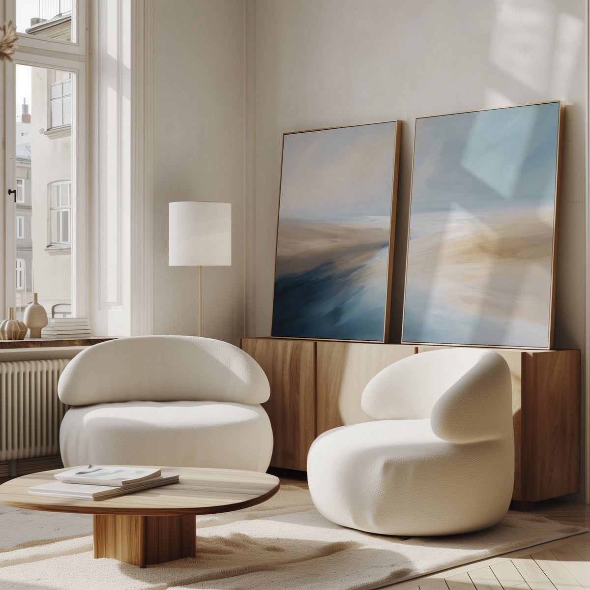 A modern living room with two curved white armchairs, a low wooden coffee table, a floor lamp, and Fluid Motion - Set Of 2 coastal wall art in blue and beige on a wooden sideboard by a large window.