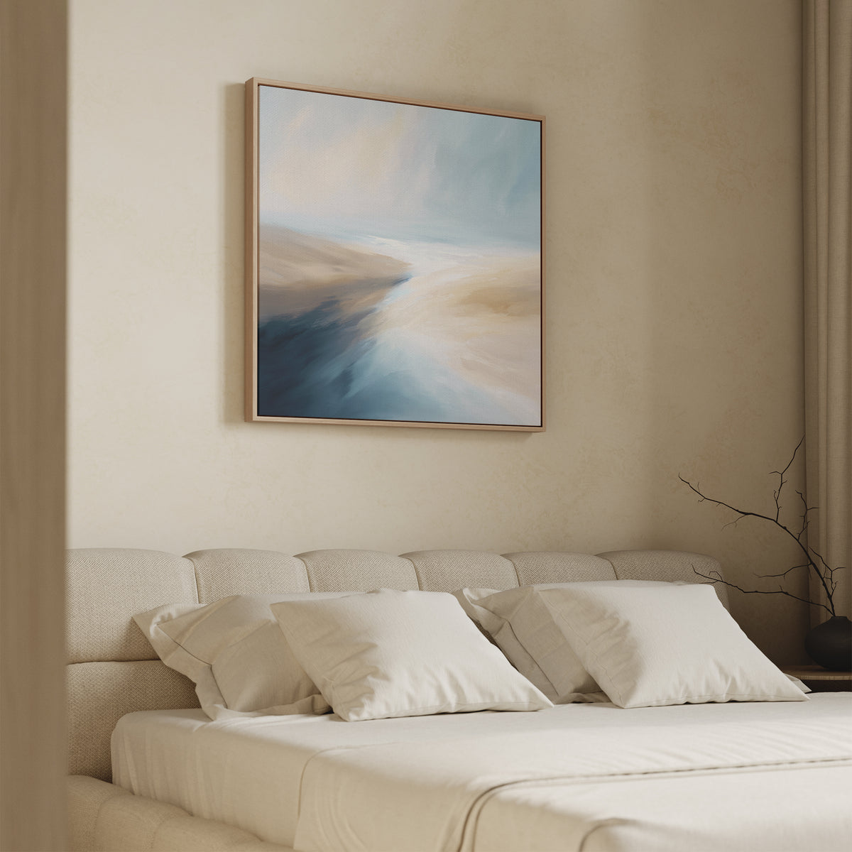 A minimal bedroom with a beige upholstered bed, four pillows, and a soft comforter. Above the bed hangs the Fluid Motion Canvas Art - Square in blue, cream, and tan, bringing a hint of coastal vibe. A bare branch sits in a vase on the nightstand.