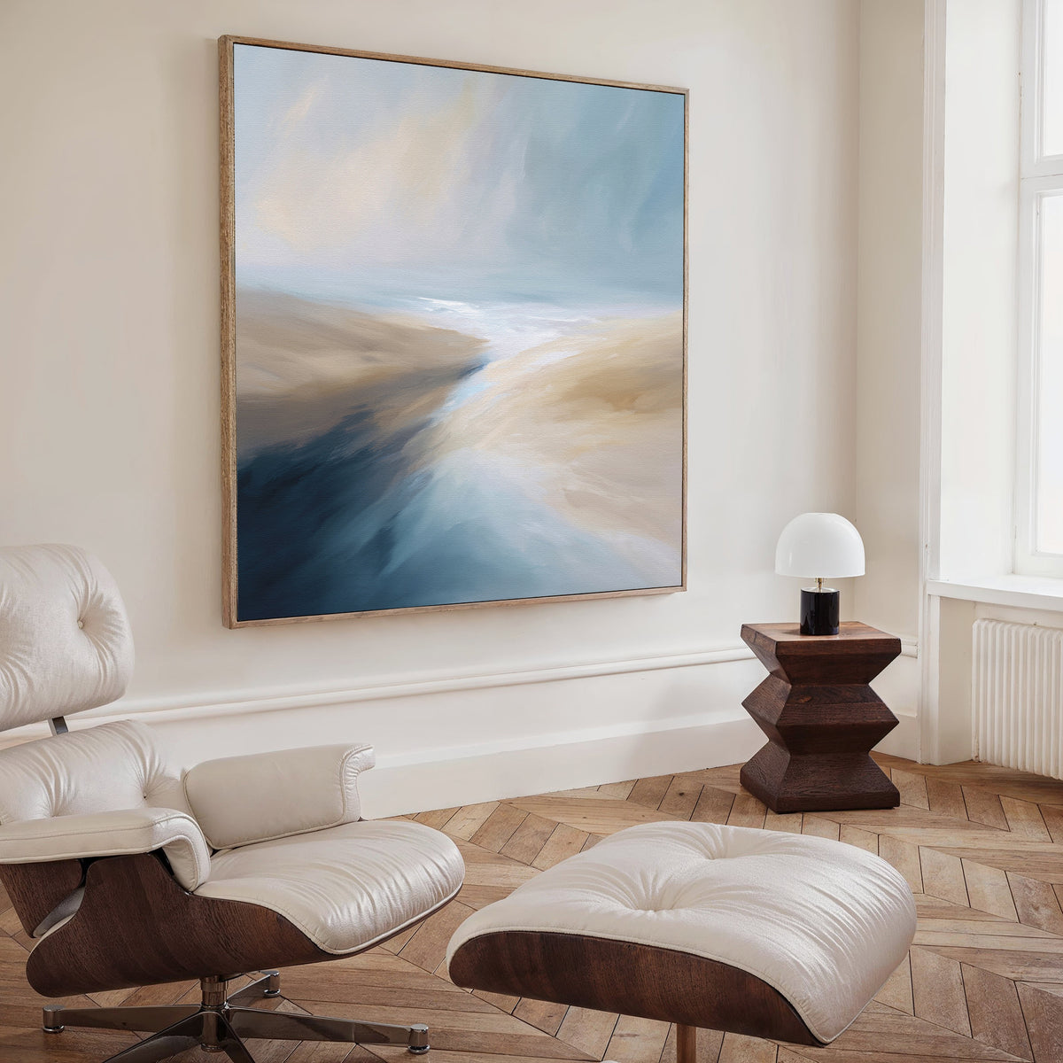 A modern living room features the Fluid Motion Canvas Art - Square in blue, beige, and white above a lounge chair and ottoman, paired with a wooden side table and white lamp by the window for a serene coastal vibe.