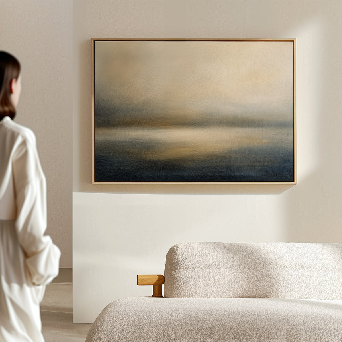 A person stands near a cream-colored sofa, admiring the Foggy Bay Impressions Canvas Art—horizontal abstract wall art in soft blue, beige, and gold tones that enhances the serene Nordic Park-inspired decor.