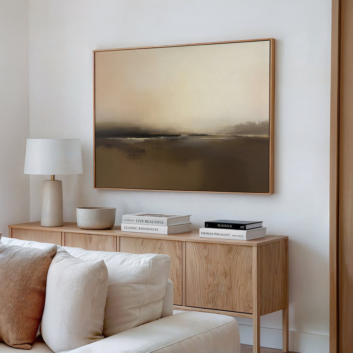 Minimalist living room featuring a light wood sideboard, neutral decor, stacked books, white lamp, and the Foggy Beach Expanse Canvas Art - Horizontal in beige and brown tones displayed on a white wall.