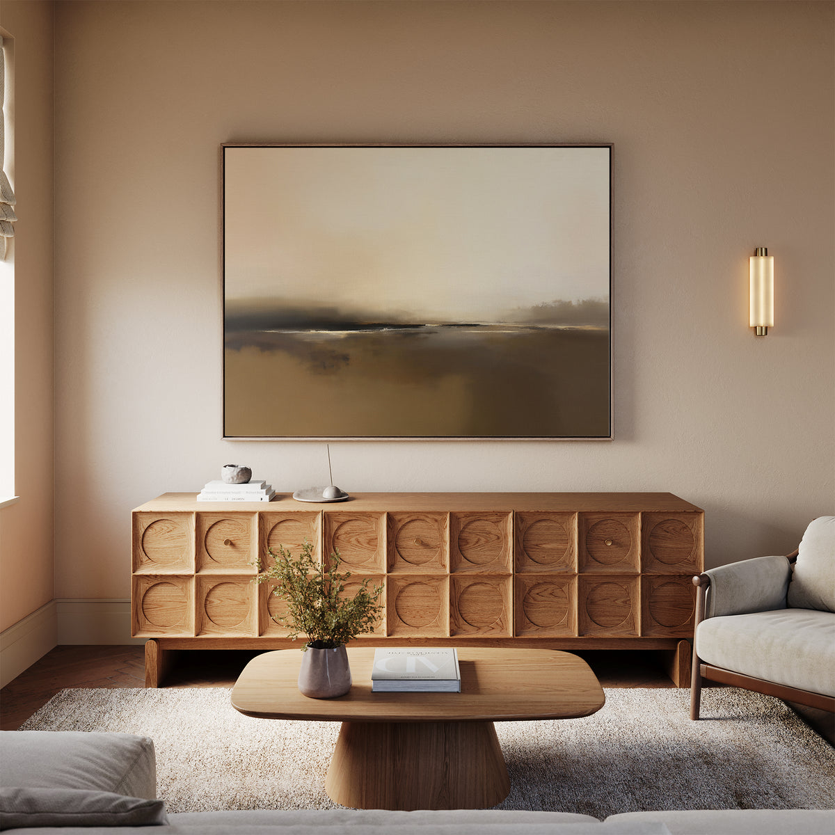 A modern living room with neutral tones features a geometric wooden sideboard, a beige sofa topped by the Foggy Beach Expanse Canvas Art - Horizontal, a wooden coffee table with books, a vase of flowers, and a soft rug on light flooring.