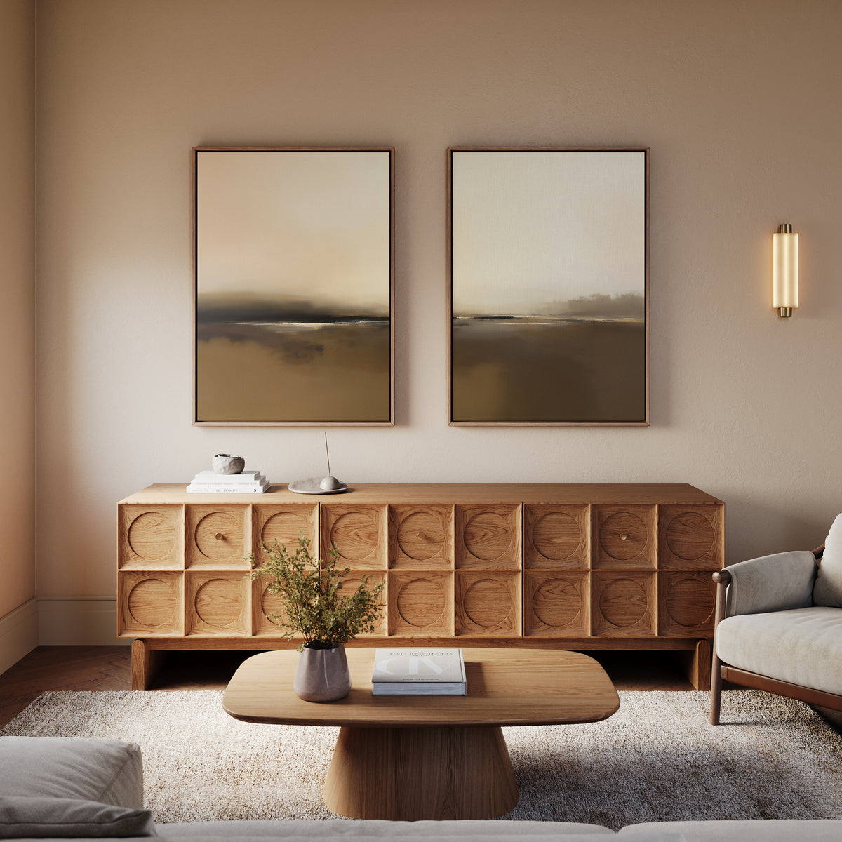 A modern living room featuring a wooden coffee table, geometric sideboard, neutral armchair, Foggy Beach Expanse - Set Of 2 artworks, sconce lighting, and a vase with greenery exudes tranquil sophistication.