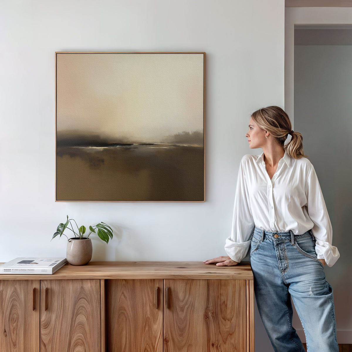 A woman in a white blouse and jeans leans against a wooden cabinet, admiring the "Foggy Beach Expanse Canvas Art - Square" on the wall. A potted plant and book atop the cabinet enhance the Nordic home decor atmosphere.