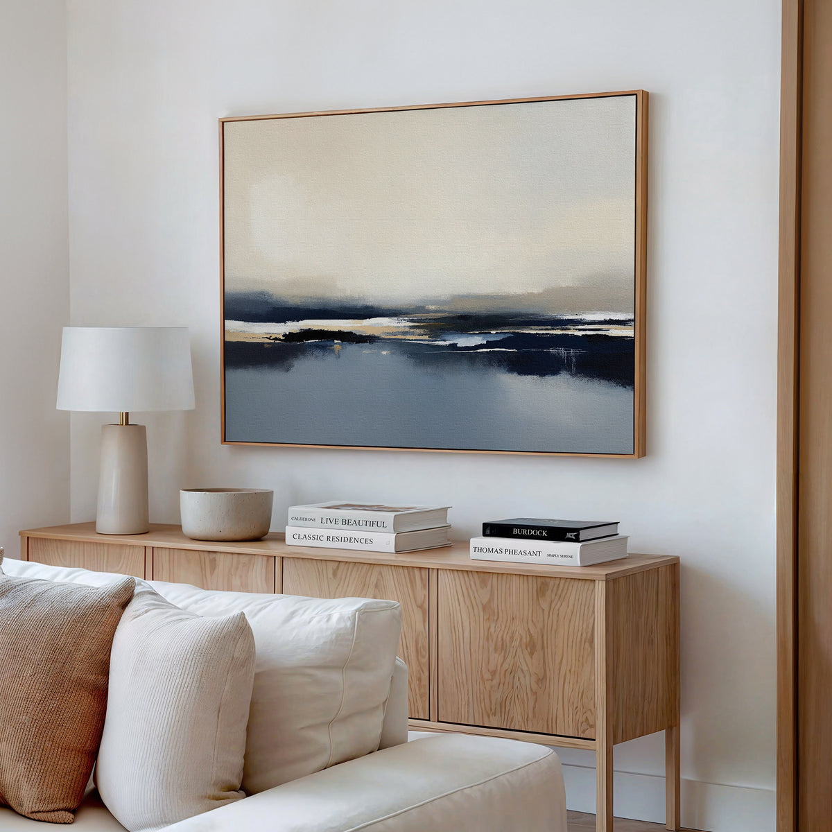 A minimalist living room with a light wood sideboard, white table lamp, stacked books, and beige sofa is completed by the "Frozen Bay Canvas Art - Horizontal," bringing serene blue and beige tones to the modern space.