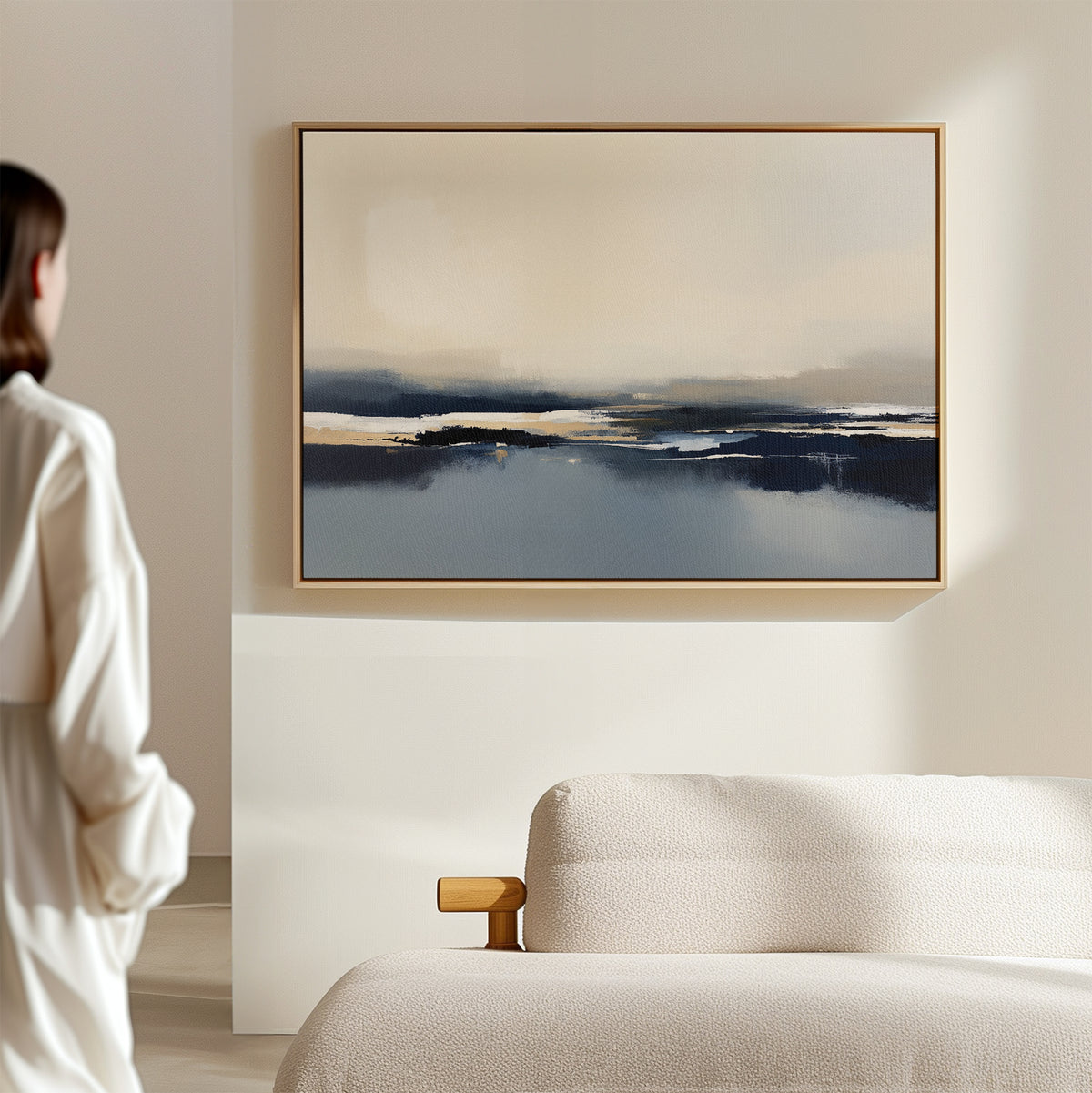 A person in a white shirt stands near a light sofa, admiring the "Frozen Bay Canvas Art - Horizontal," featuring blue, beige, and white winter water tones on a pale wall in a modern living room.