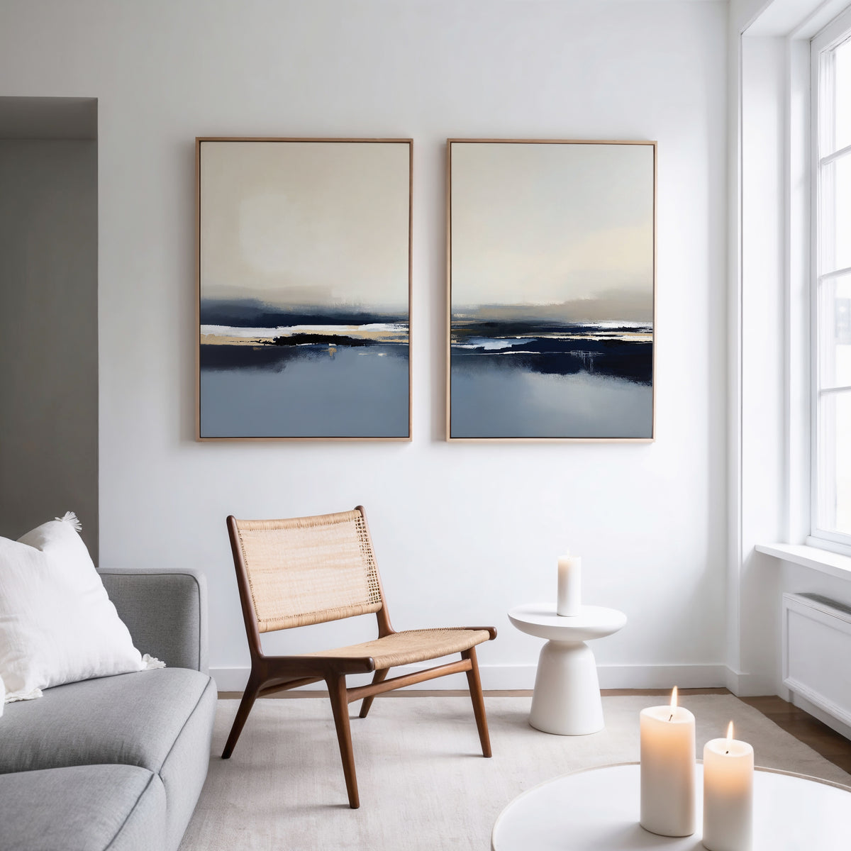 A minimalist living room with a woven chair, side table with a lit candle, gray sofa, and the Frozen Bay - Set Of 2 landscape prints on the wall, all illuminated by natural light from a large window.