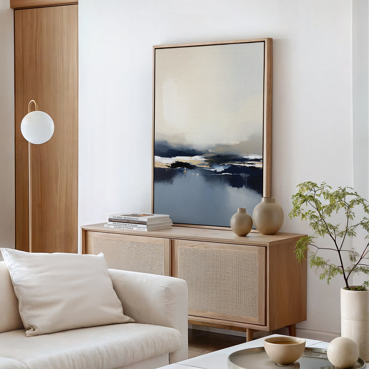 A modern living room featuring light wood furniture, Frozen Bay Canvas Art - Vertical above a sideboard, ceramic vases, stacked books, a round lamp, green potted plant, and a cream-colored sofa with cushions.