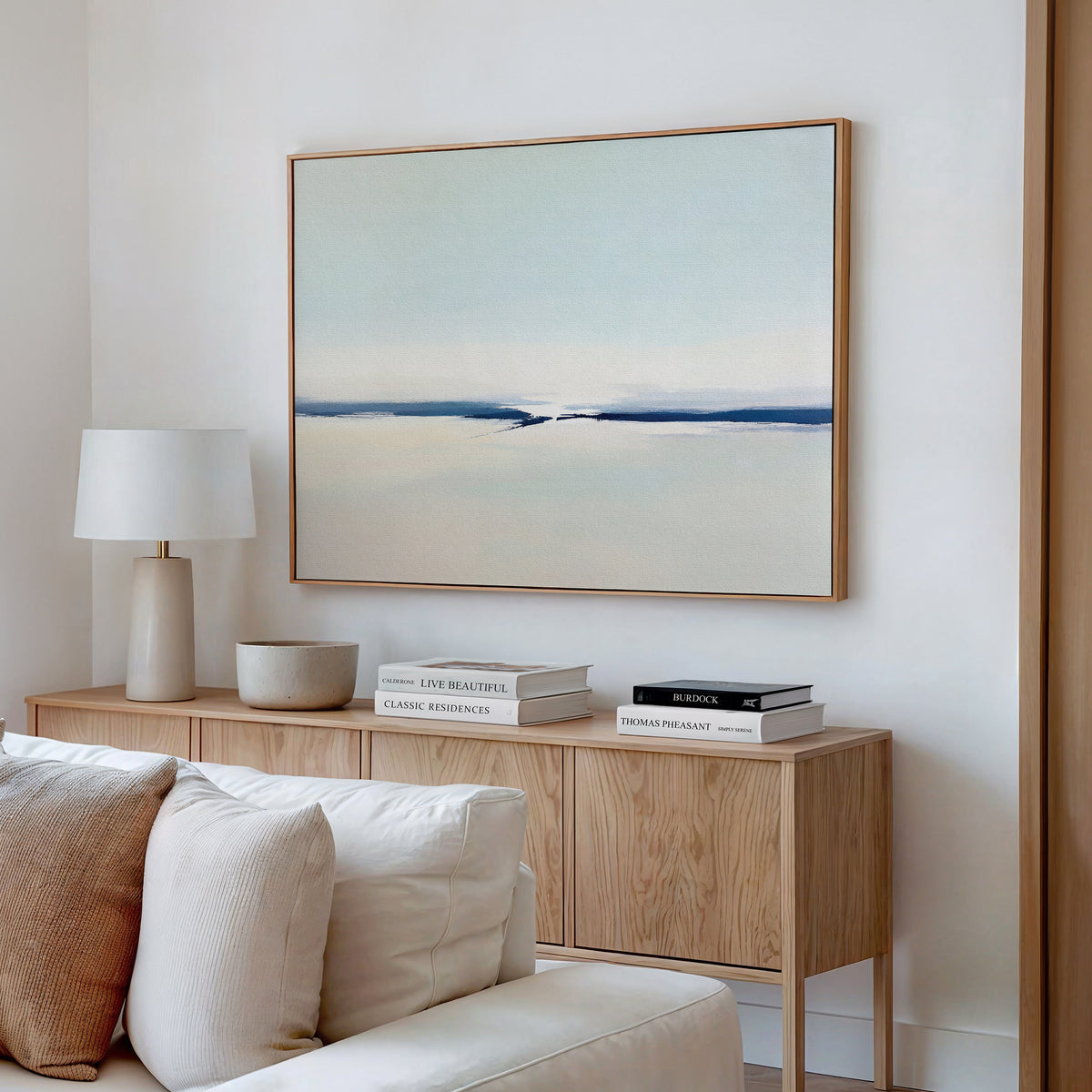 A minimalist living room features a light wood console with books, a lamp, and decor, a cream sofa, and the Frozen Channel Canvas Art - Horizontal in soft blue and white tones on a white wall.
