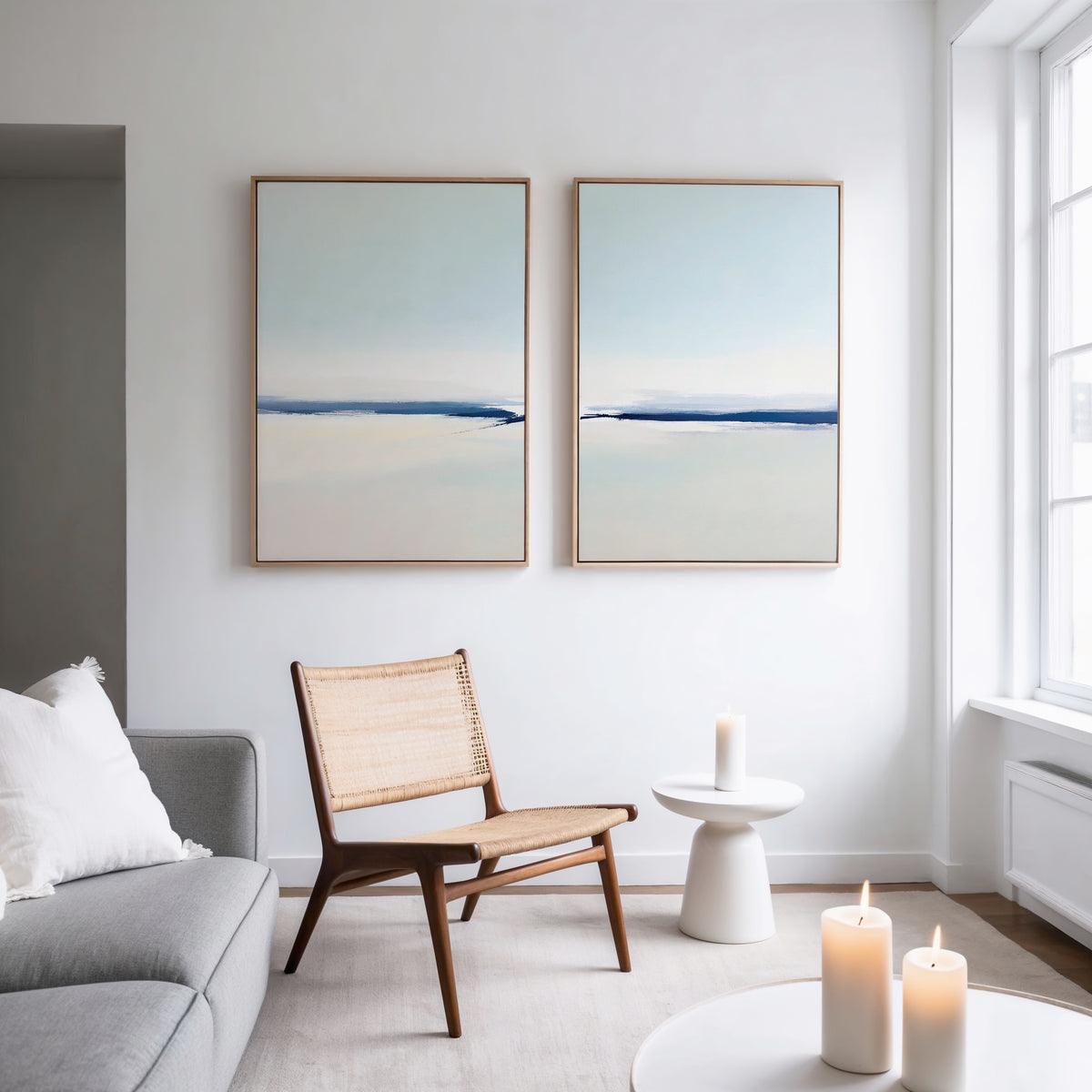 A minimalist living room features a gray sofa, woven chair, white side table with candle, and the Frozen Channel - Set of 2 art prints as serene wall art. Large windows fill the space with light for a modern decor vibe.