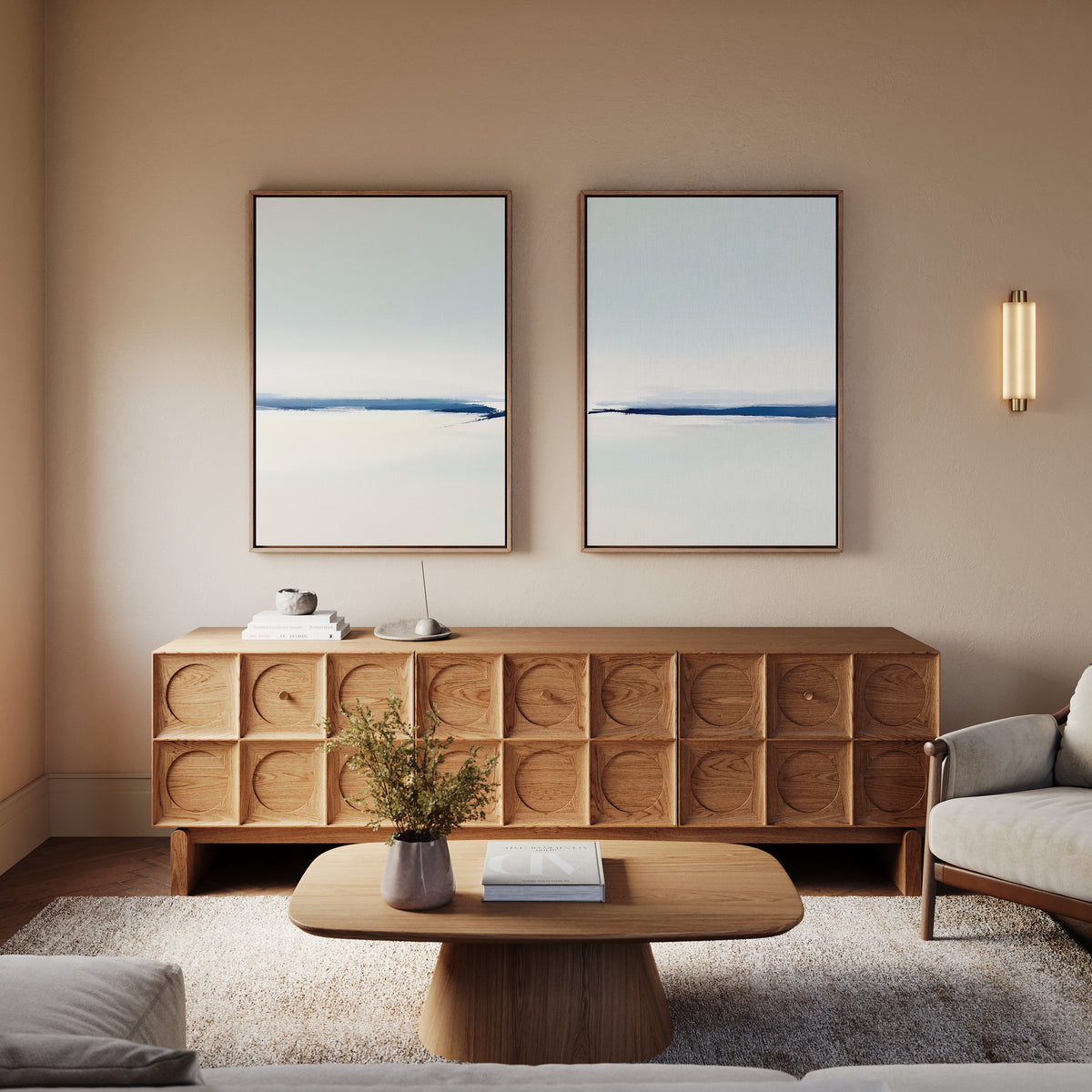 A serene modern living room features the Frozen Channel - Set Of 2 wall art above a wooden sideboard, a low coffee table with books and a plant, a neutral armchair, and sconce lighting for warmth.