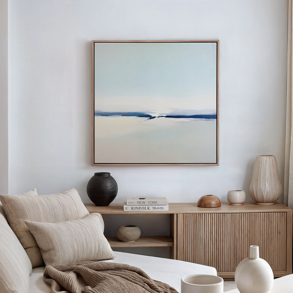 A minimalist living room with a beige sofa, neutral pillows, a wooden sideboard topped with decorative vases and books, and the Frozen Channel Canvas Art - Square in blue and white tones as striking wall art above.