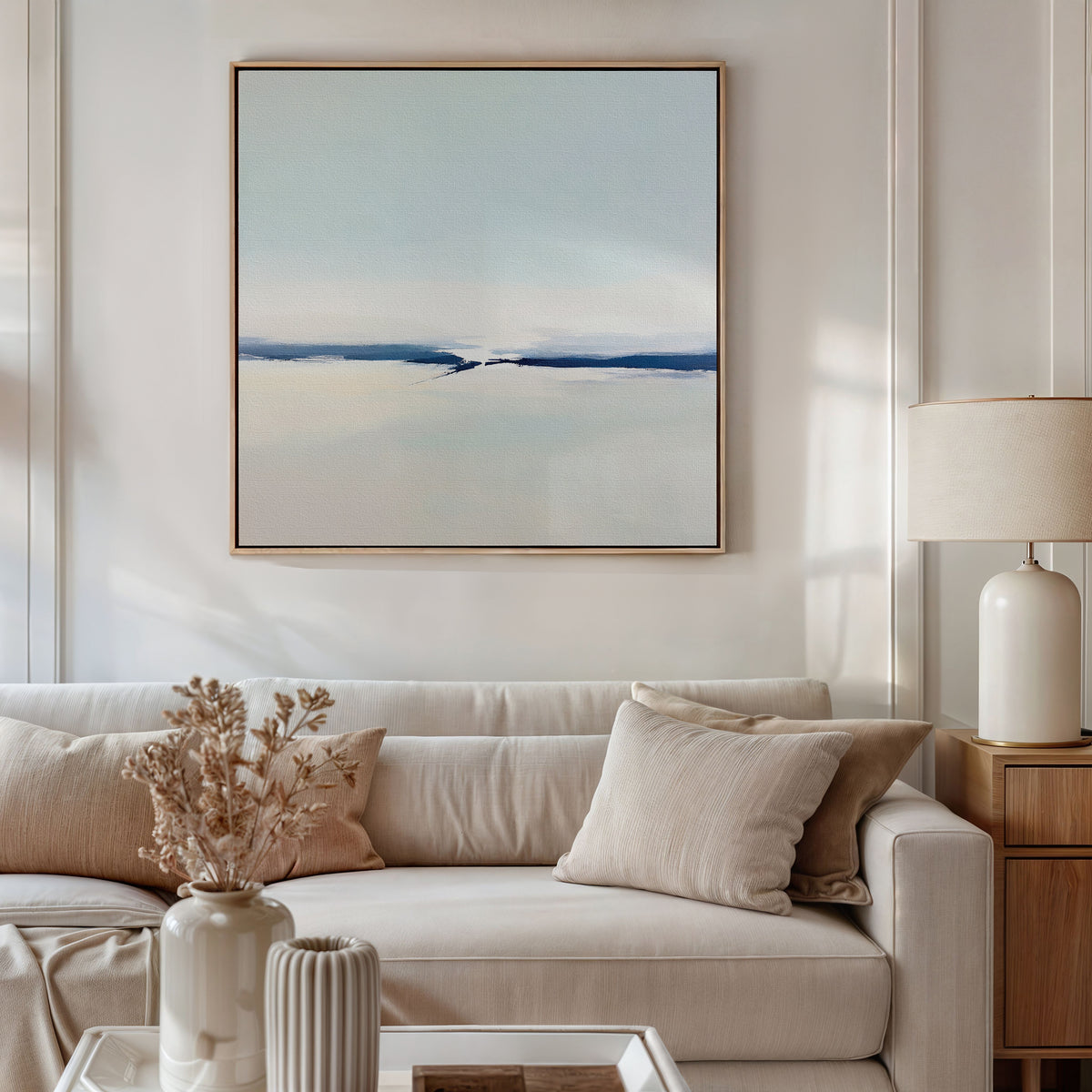 A modern living room features a beige sofa, neutral pillows, a wooden side table with a ceramic lamp, and the Frozen Channel Canvas Art - Square in soft blue and white tones above the couch. Dried flowers on the coffee table complete the look.