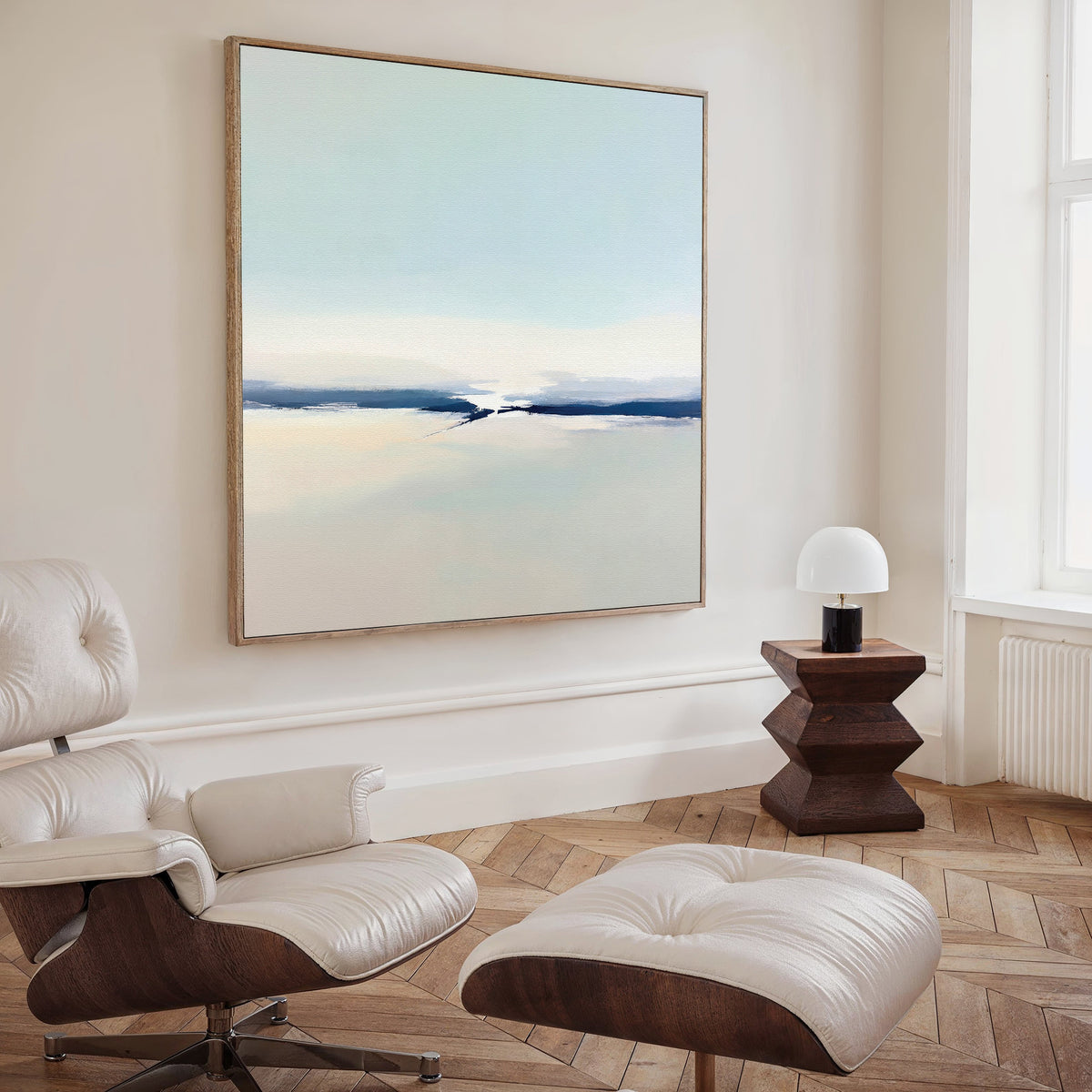 A minimalist living room with a white leather lounge chair, ottoman, wooden side table, and modern decor featuring the Frozen Channel Canvas Art - Square in soft blue and beige tones on the wall.