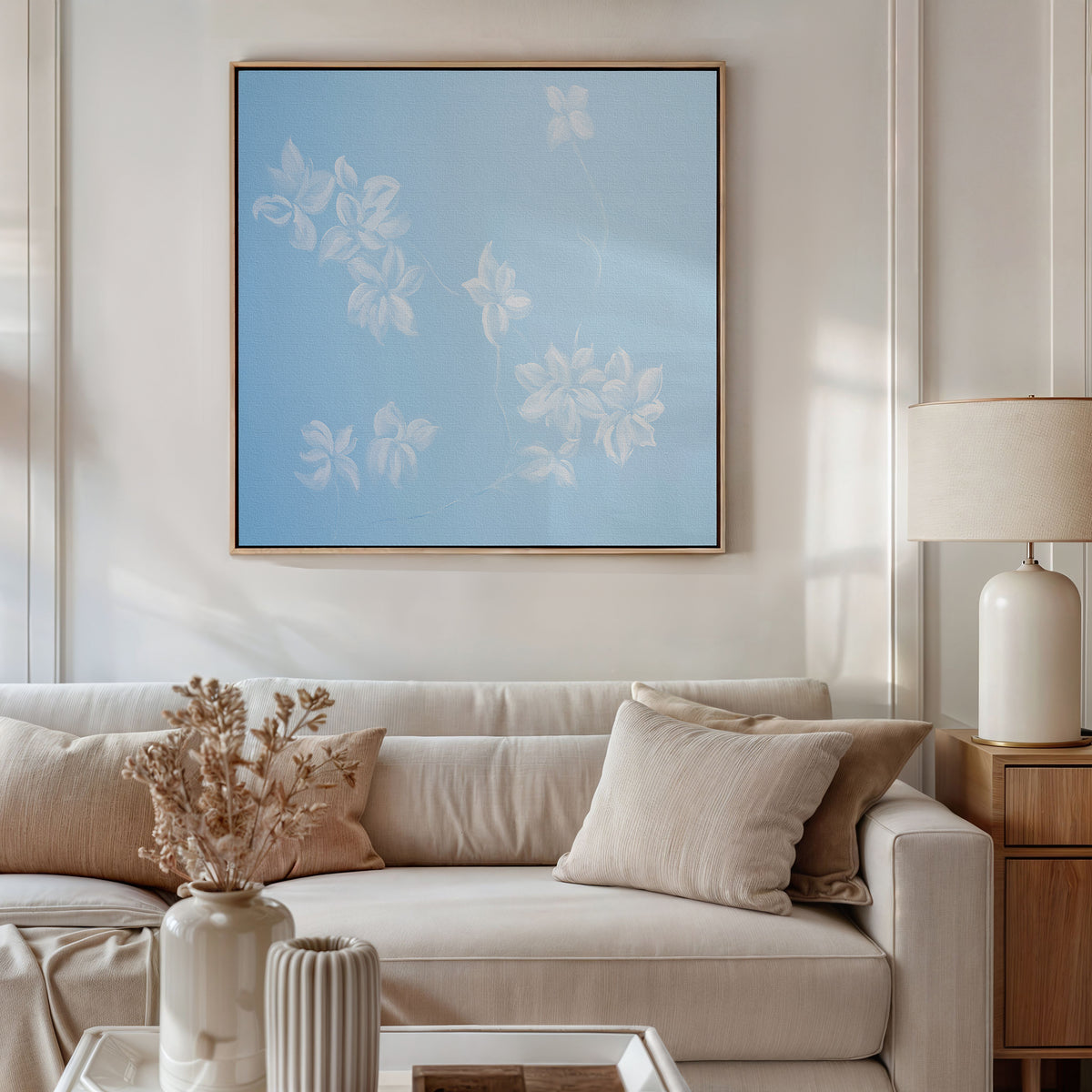 A modern living room with a beige sofa, neutral pillows, a side table with a lamp, and dried flowers in a vase. Above the sofa hangs the Gentle Blossoms Canvas Art - Square, featuring elegant white floral designs on a blue background.
