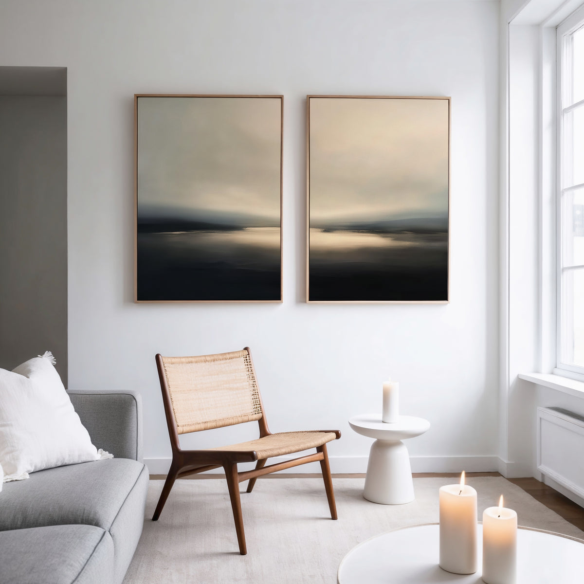 A modern, minimalist living room with a gray sofa, woven chair, “Golden Fog Horizon - Set Of 2” wall art above, a small white side table, and three lit candles for a calm atmosphere.