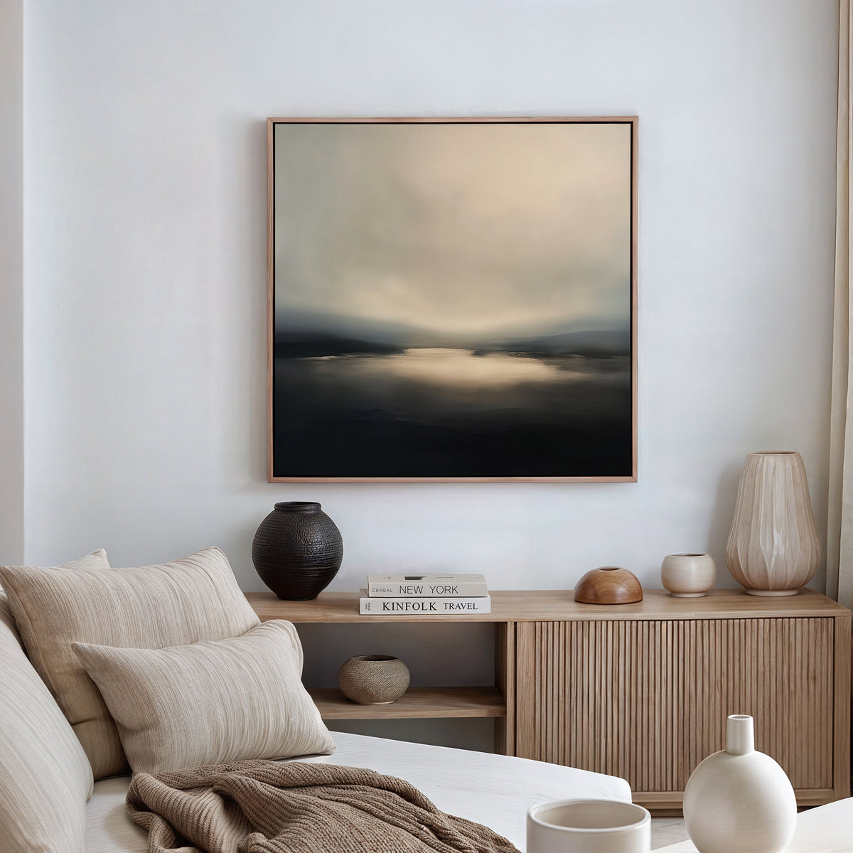 A minimalist living room featuring a light sofa, neutral pillows, a wooden sideboard topped with ceramic vases and books, and the Golden Fog Horizon Canvas Art - Square in soft, muted tones displayed above the sideboard.