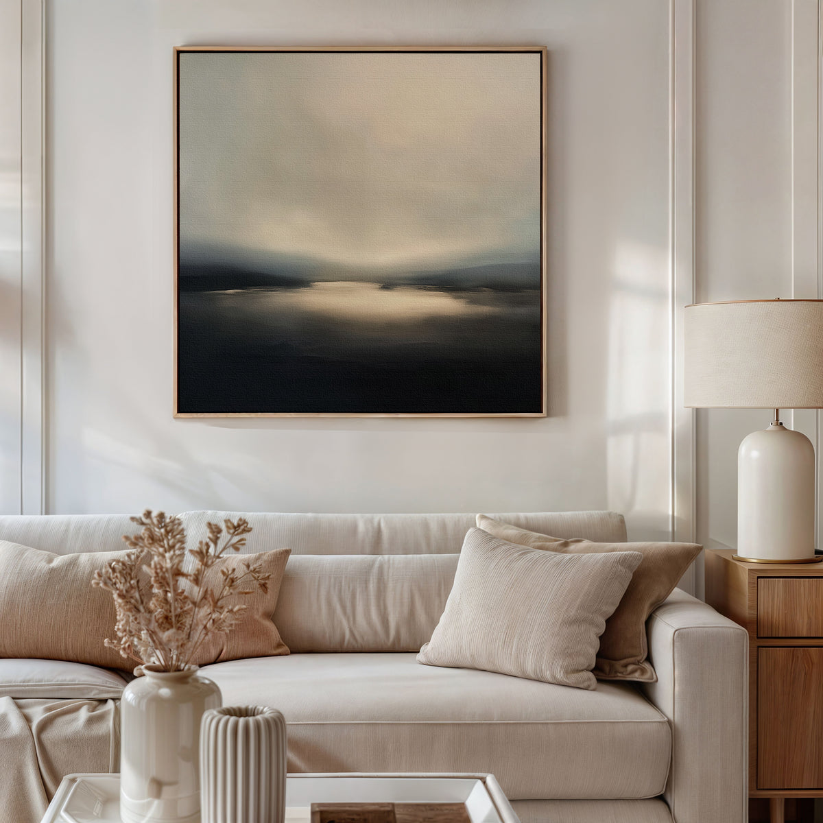 A modern living room features a beige sofa, neutral pillows, and a wooden side table with a white lamp. Above the sofa hangs the Golden Fog Horizon Canvas Art - Square. Dried flowers in a vase sit on the coffee table.