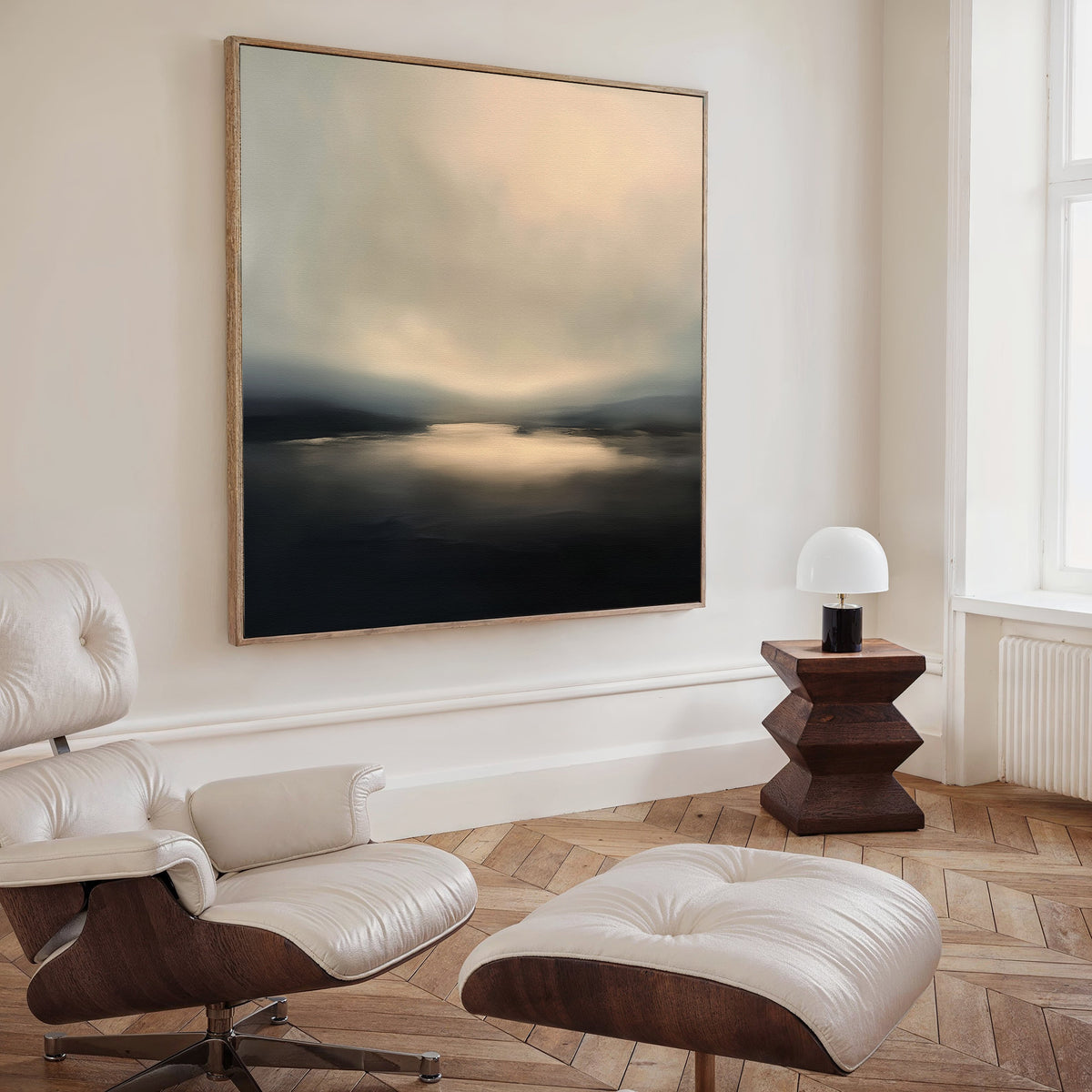 A modern living room with a white leather lounge chair and ottoman, a wooden side table with a small white lamp, and the Golden Fog Horizon Canvas Art - Square on the wall. Sunlight pours in through a tall window.