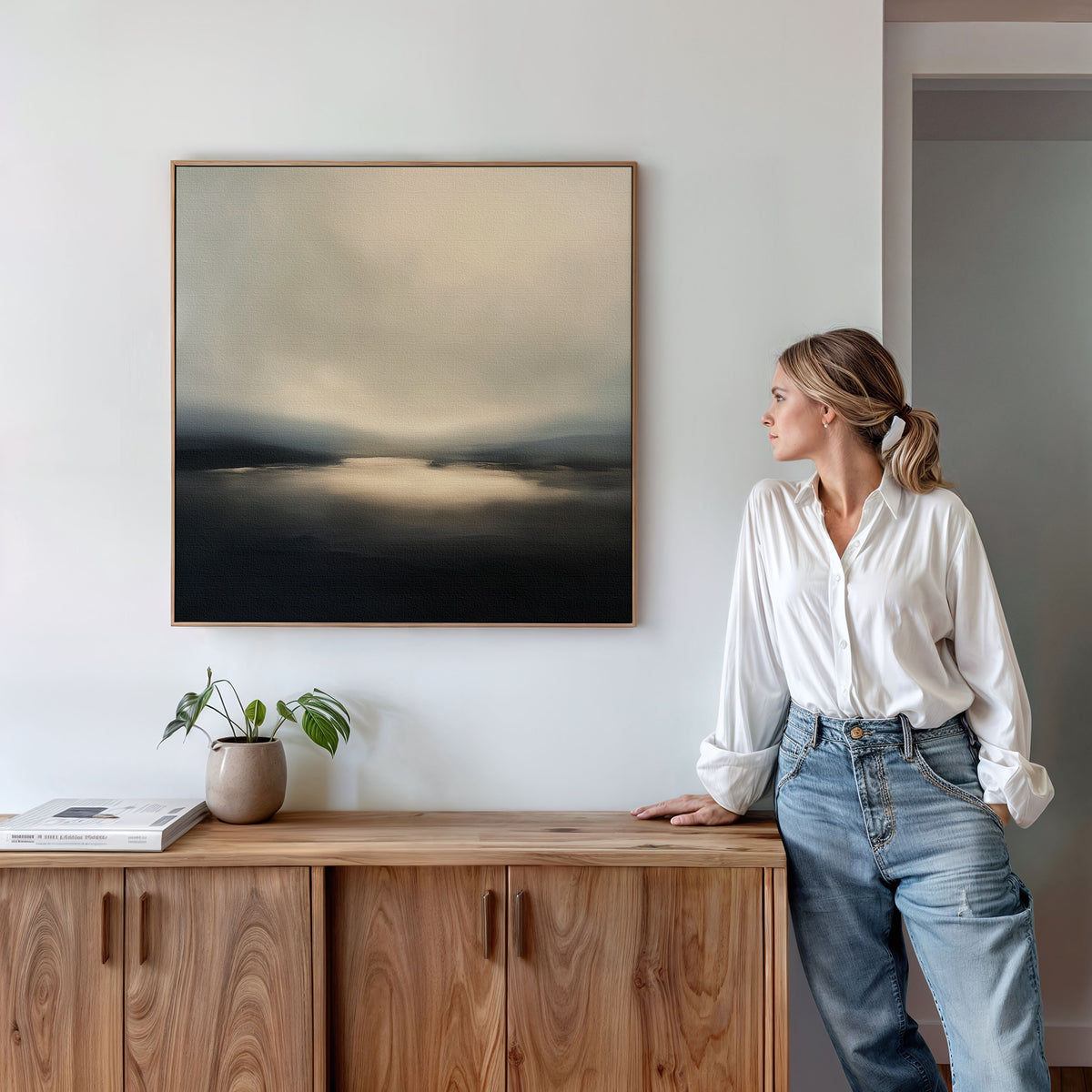 A woman in a white blouse and jeans stands by a wooden cabinet, admiring the Golden Fog Horizon Canvas Art - Square on the wall; a small potted plant and books sit neatly atop the cabinet.
