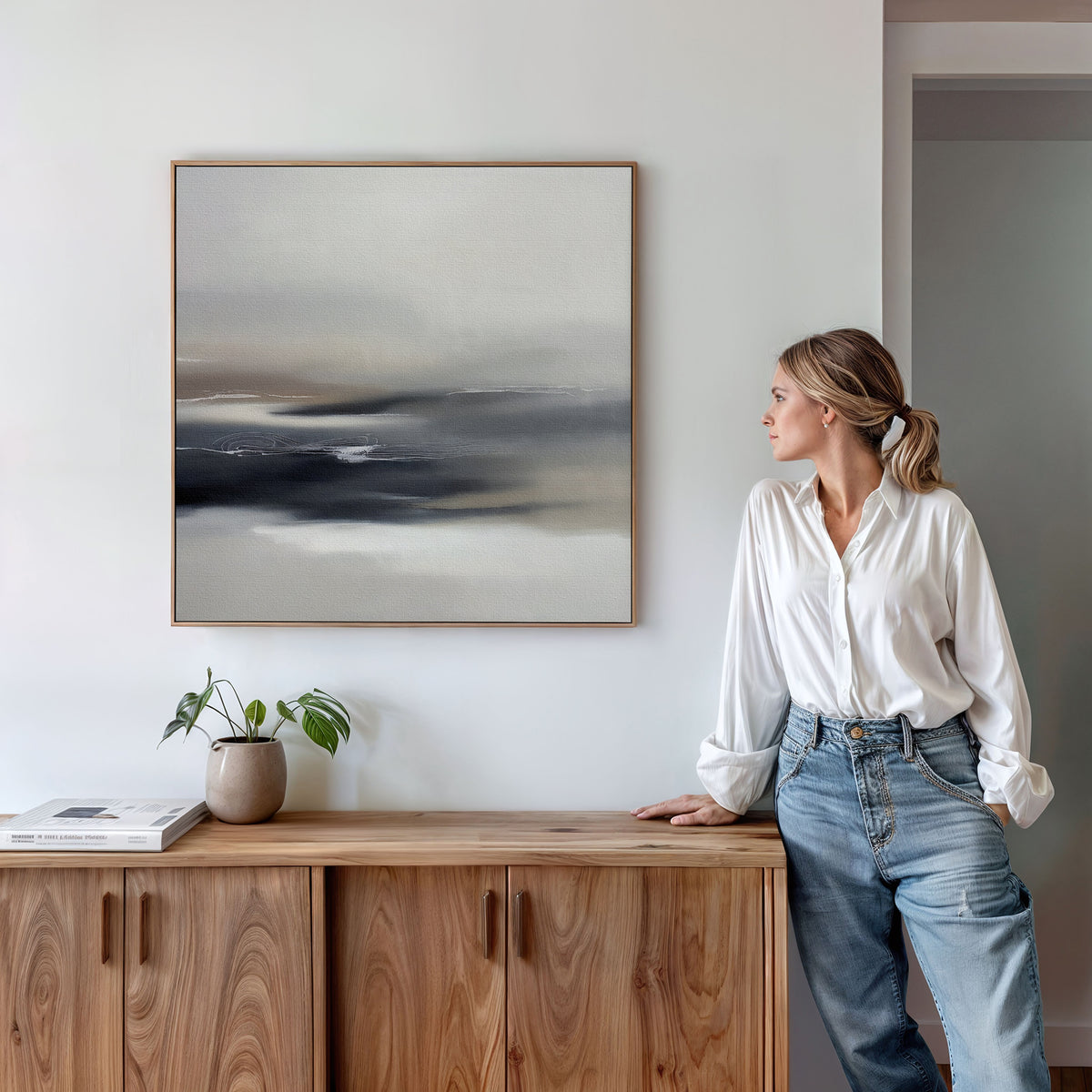 A woman in a white blouse and jeans leans against a wooden cabinet, gazing at the Grey Ripples Canvas Art - Square on a white wall. A potted plant and books rest on the cabinet, enhancing this modern living room scene.