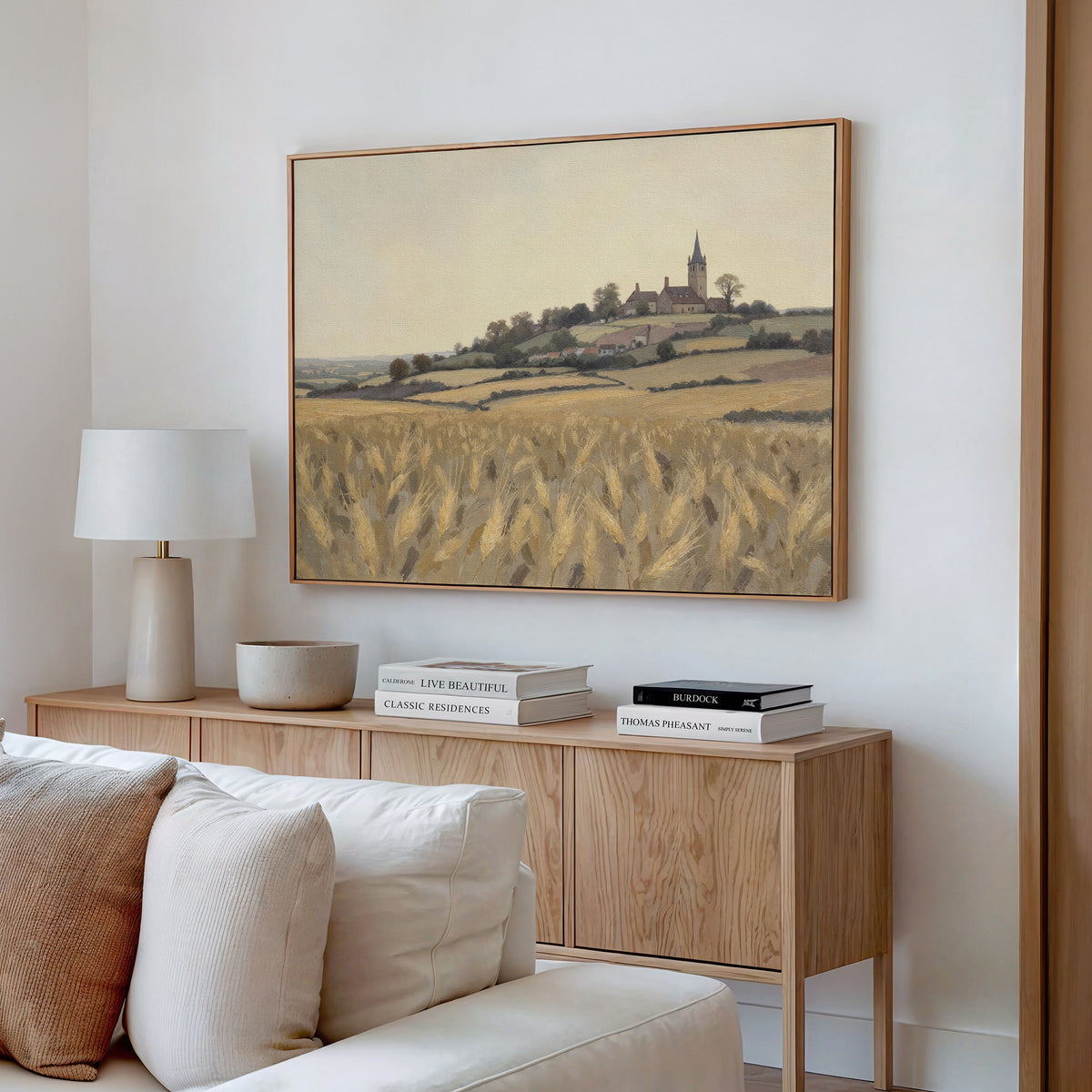 A cozy living room with a wooden sideboard, books, a lamp, and ceramics, featuring rustic wall art—the Harvest Chapel Canvas Art (Horizontal), depicting a church and village on a hill above a golden wheat field.
