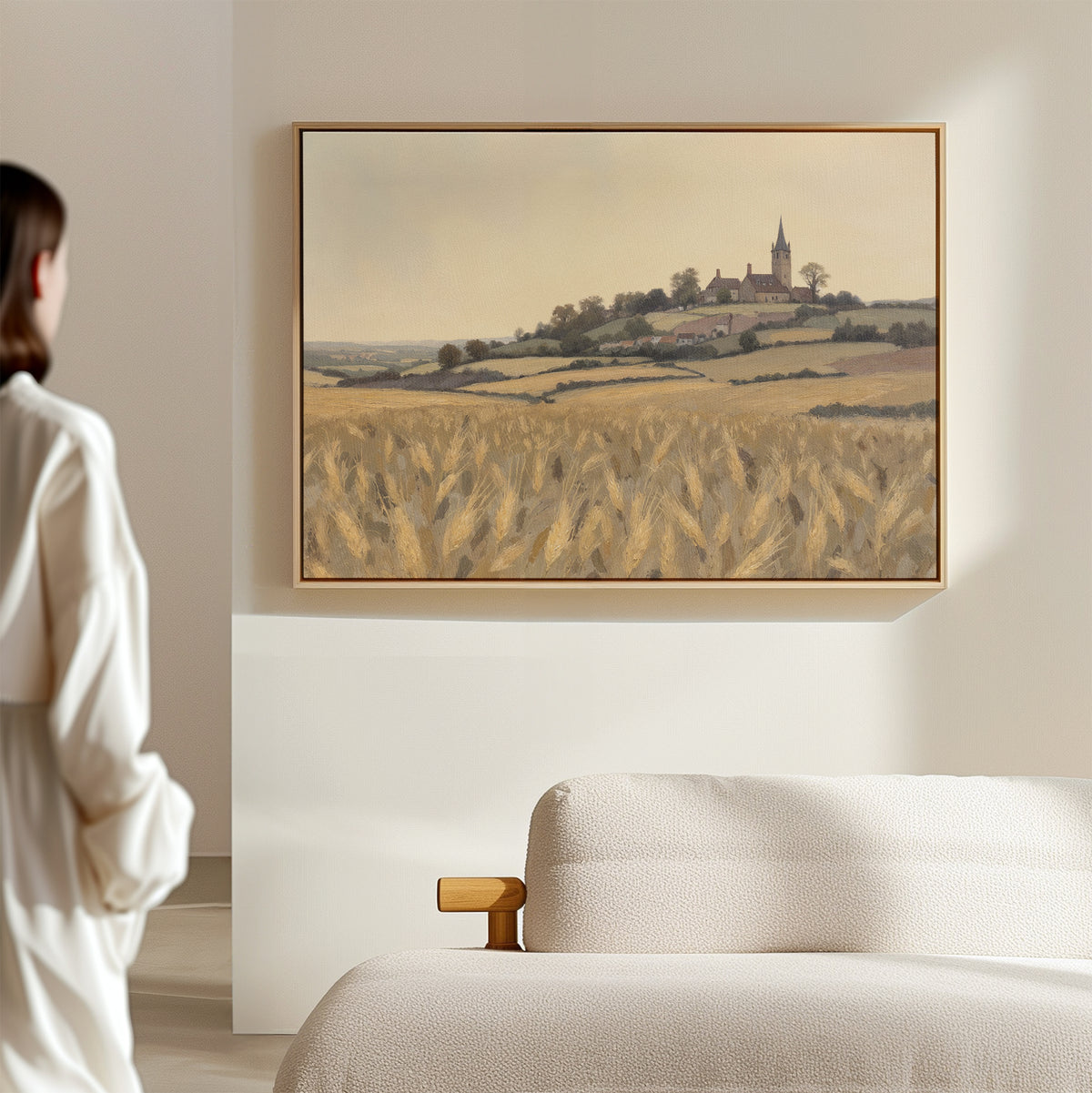 A woman stands in a softly lit room, admiring the Harvest Chapel Canvas Art - Horizontal, featuring golden fields and a distant village with a church steeple, displayed above a light-colored sofa.