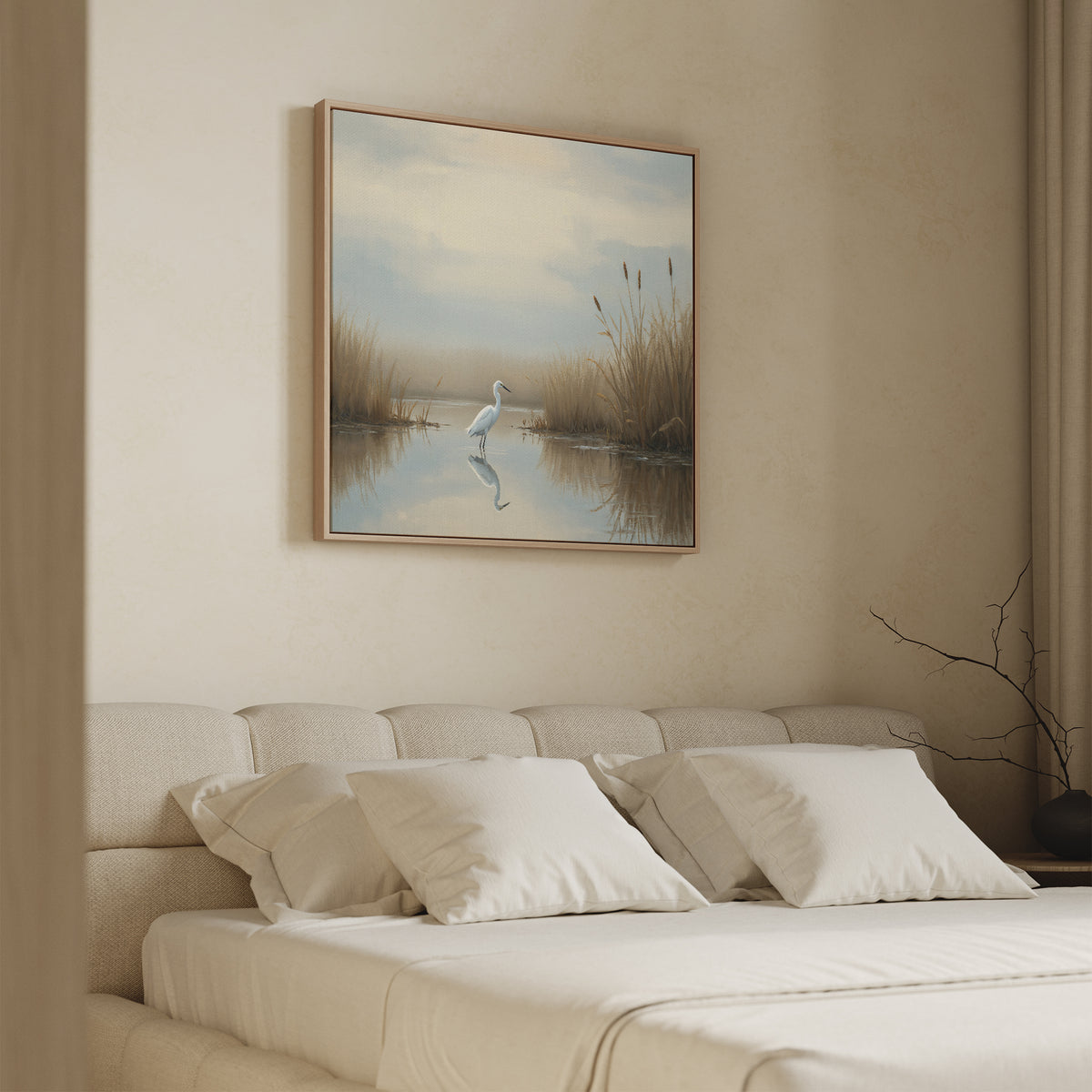 A beige bedroom features the Heron's Morning Canvas Art - Square above the bed with white pillows, showing a heron standing in water under a cloudy sky. A bare branch in a vase accents the bedside table.